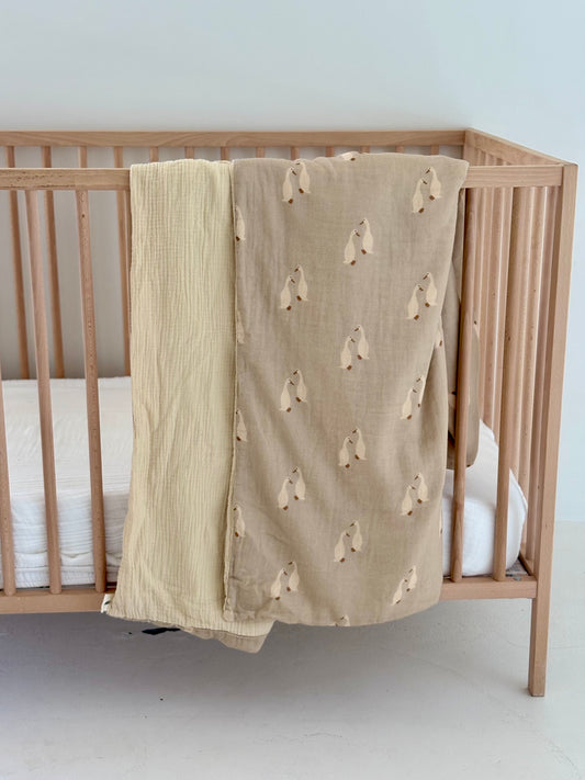 A wooden baby crib with a white mattress features two blankets: a solid light yellow muslin quilt and forever french baby's beige Muslin Quilt | Silly Goose, draped over the side—perfect for adding charm to any nursery. The background is minimal.