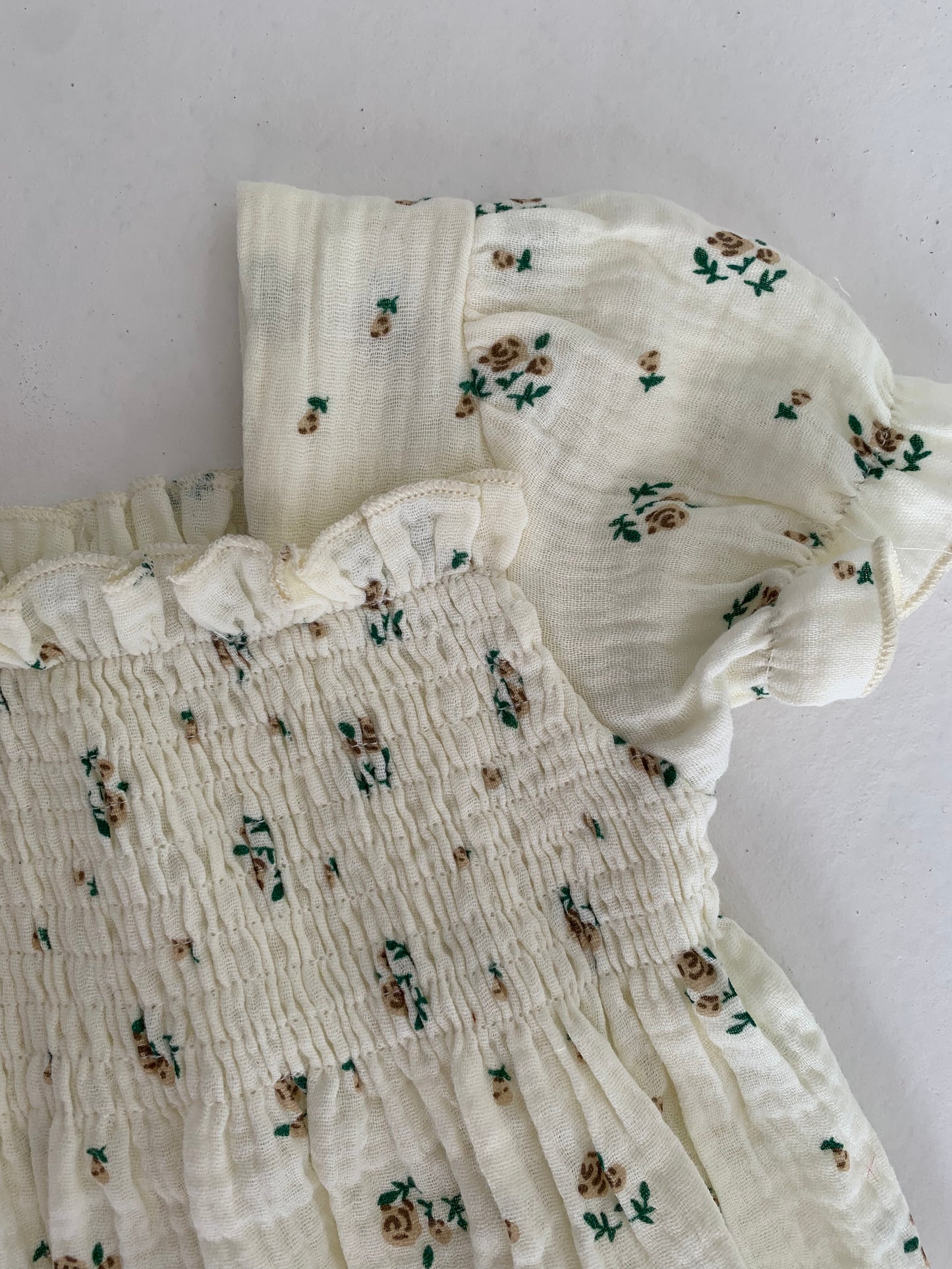 A close-up of the Garden Days Ivory Floral Romper from Forever French Baby showcases its light cream fabric with a smocked bodice adorned with small green and brown floral patterns. The romper features gathered short sleeves and a subtle ruffle trim at the top, embodying Forever French Baby's delicate charm.