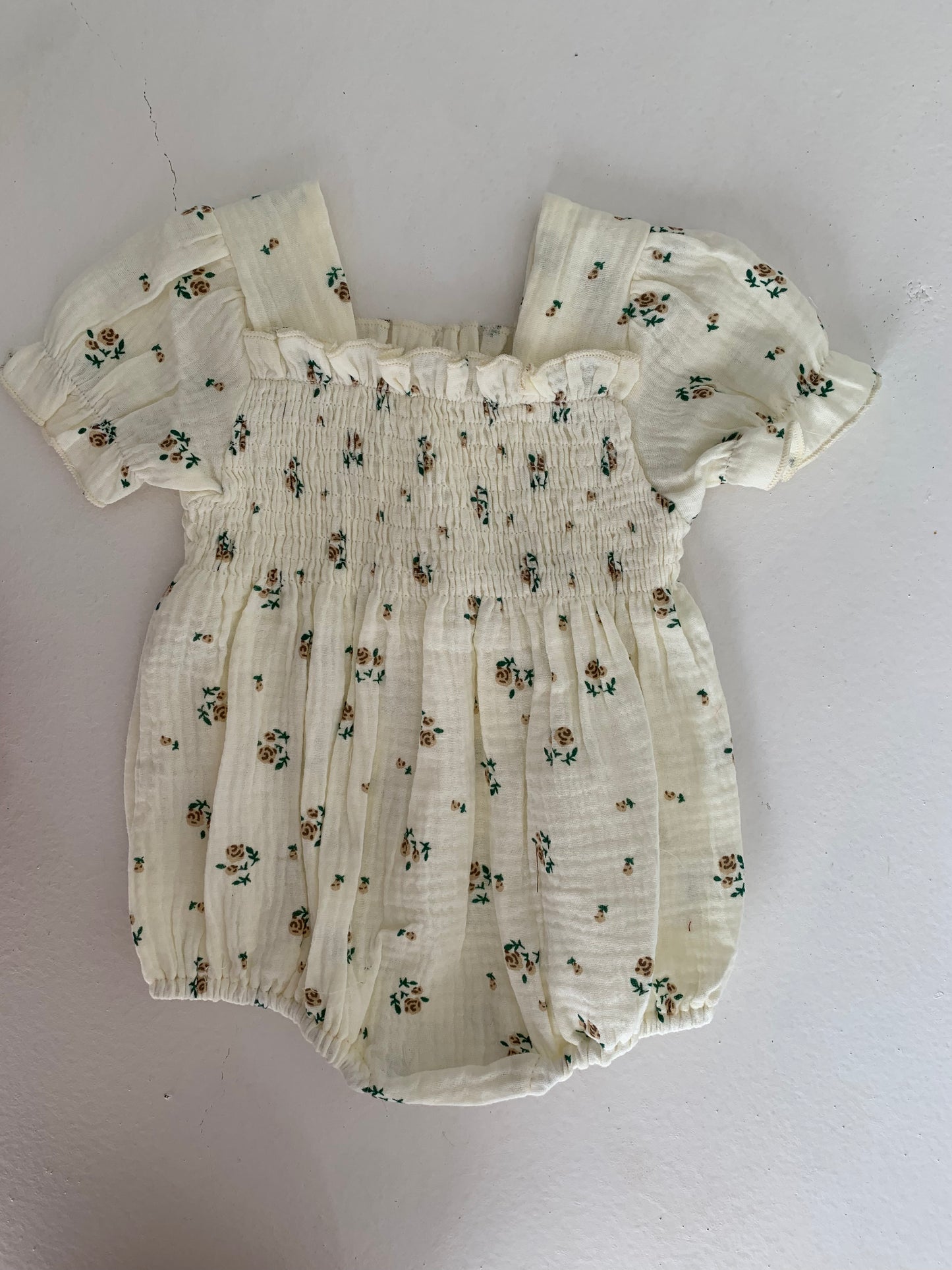 The Garden Days | Ivory Floral Romper from forever french baby features light-colored fabric with a floral pattern. It has short, puffed sleeves, smocked detailing on the chest, and elastic at the leg openings. The muslin fabric gives it a light, textured appearance.