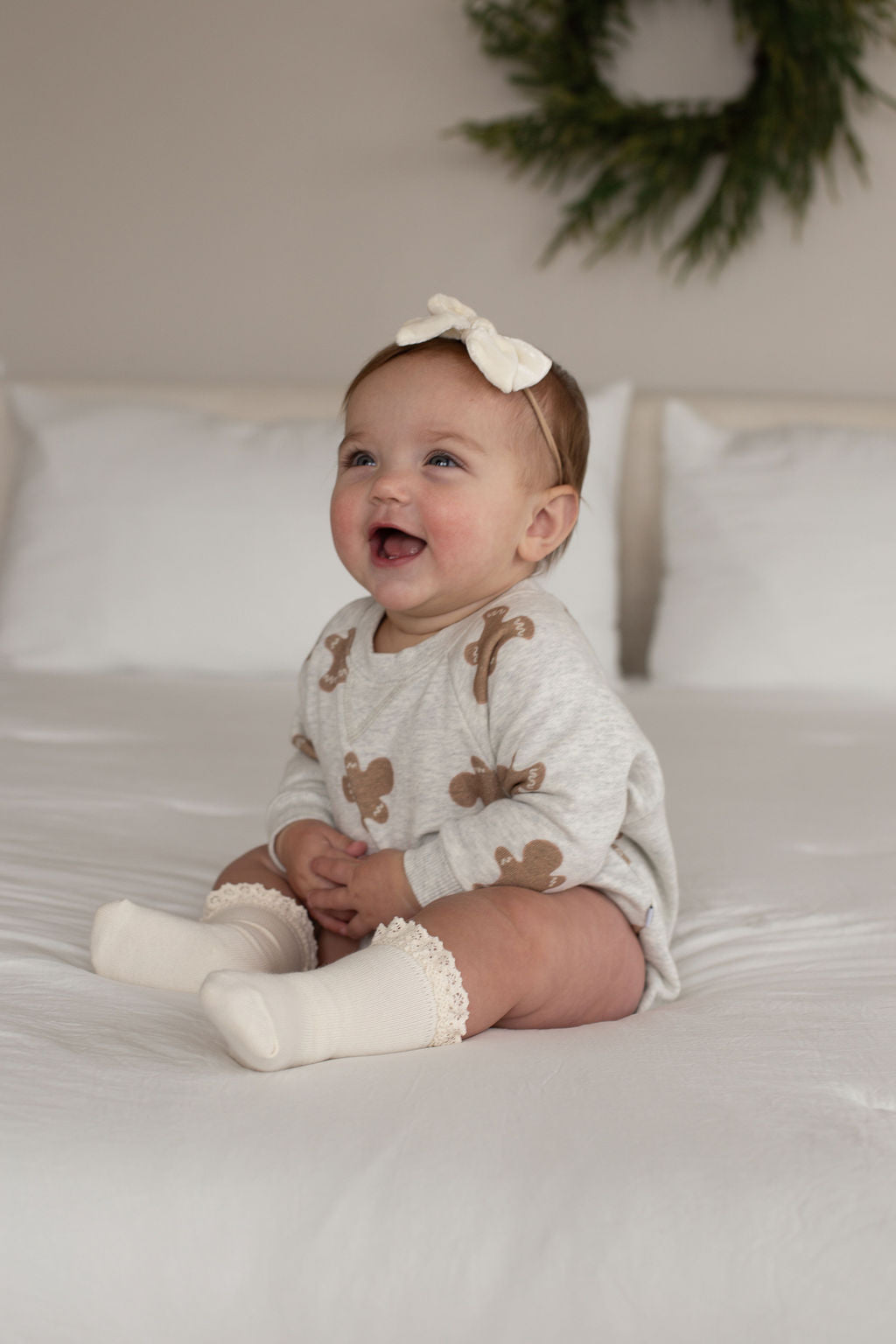 A smiling baby in the forever french baby Sweatshirt Romper | Gingerbread 2.0, a cream headband, and cream socks sits on a white bed with white pillows and a green wreath on the wall behind.