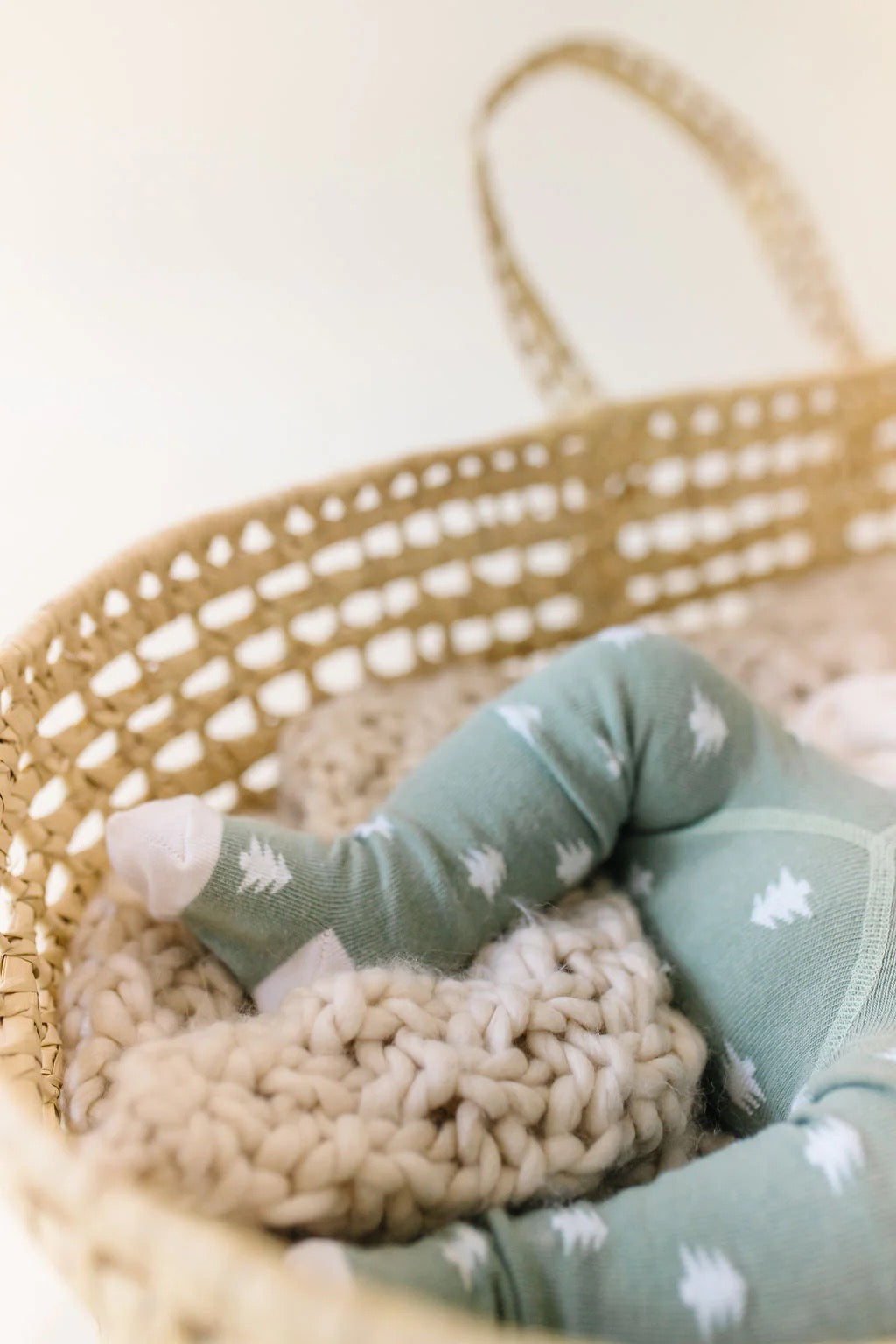 A close-up of a baby in Little Millers Pine Tights, featuring white tree patterns on sage green OEKO-TEX certified fabric. Only the legs and feet are visible, resting in a woven basket on a chunky knit blanket.