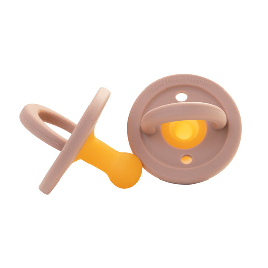 Two modern pacifiers from forever french baby's Mahogany Rose collection are shown in the image. One silicone pacifier stands upright, featuring its bulb-shaped yellow nipple and round beige guard, while the other lies flat, showcasing the back side of the guard—perfect baby essentials for your little one.