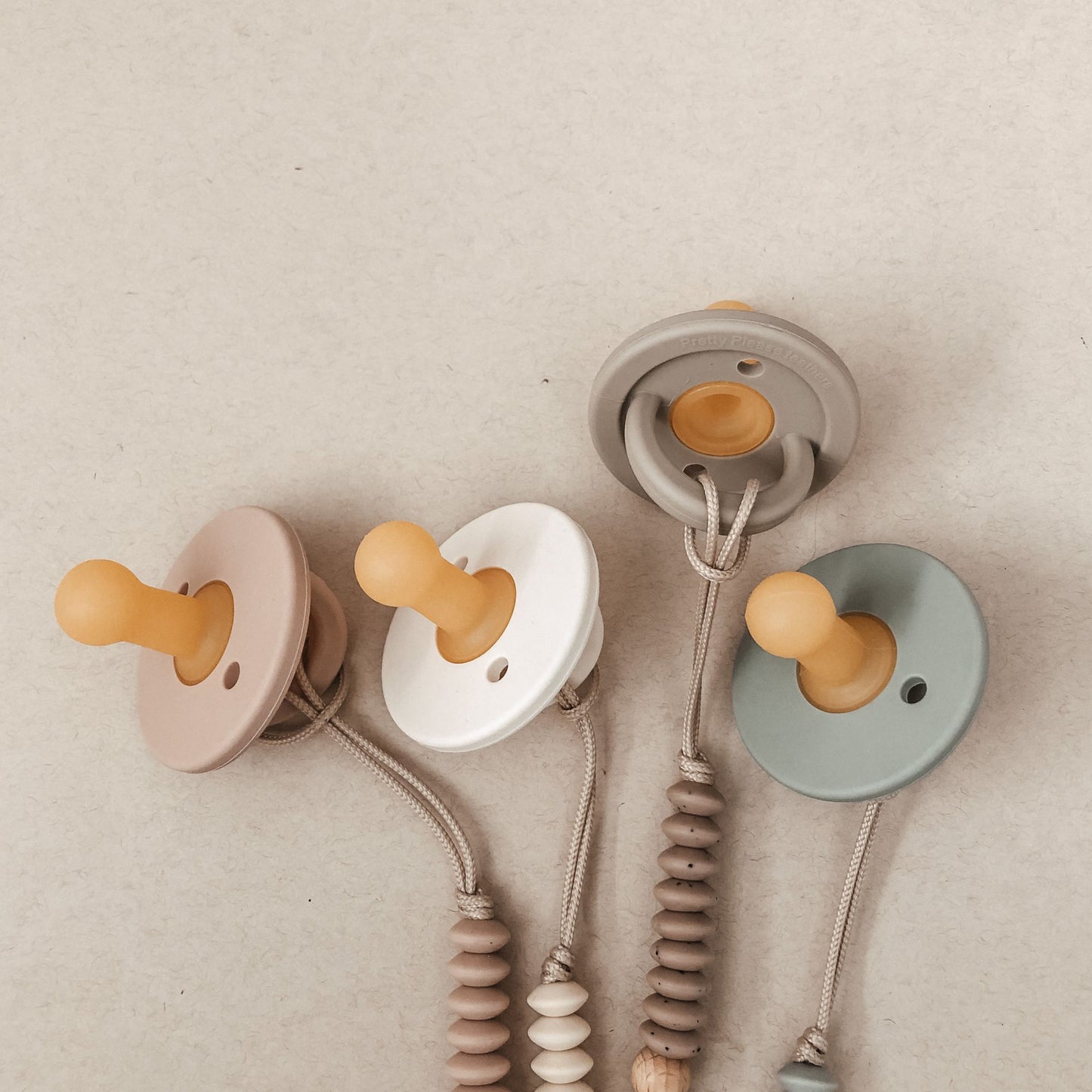 Four baby essentials, the Modern Pacifiers from forever french baby, are laid out on a beige surface. These silicone pacifiers come in pastel colors: light pink, white, gray, and teal. Each features a rounded nipple and a circular shield. The attached beaded clips have coordinated bead designs.