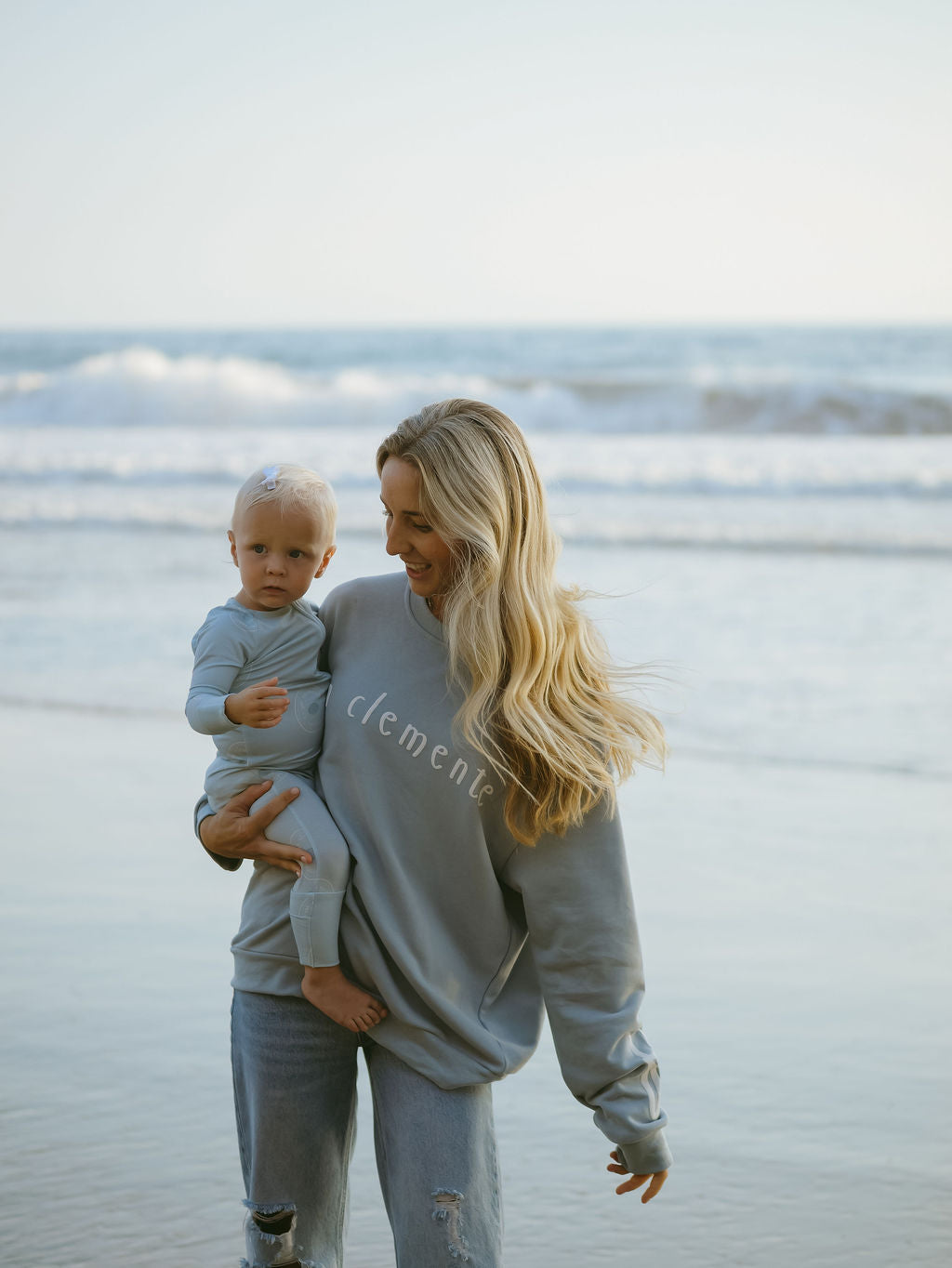A woman with long blonde hair wears the forever french baby Adult Sweatshirt | San Clemente and ripped jeans, holding a baby in matching gray attire on a beach with gentle waves behind them.