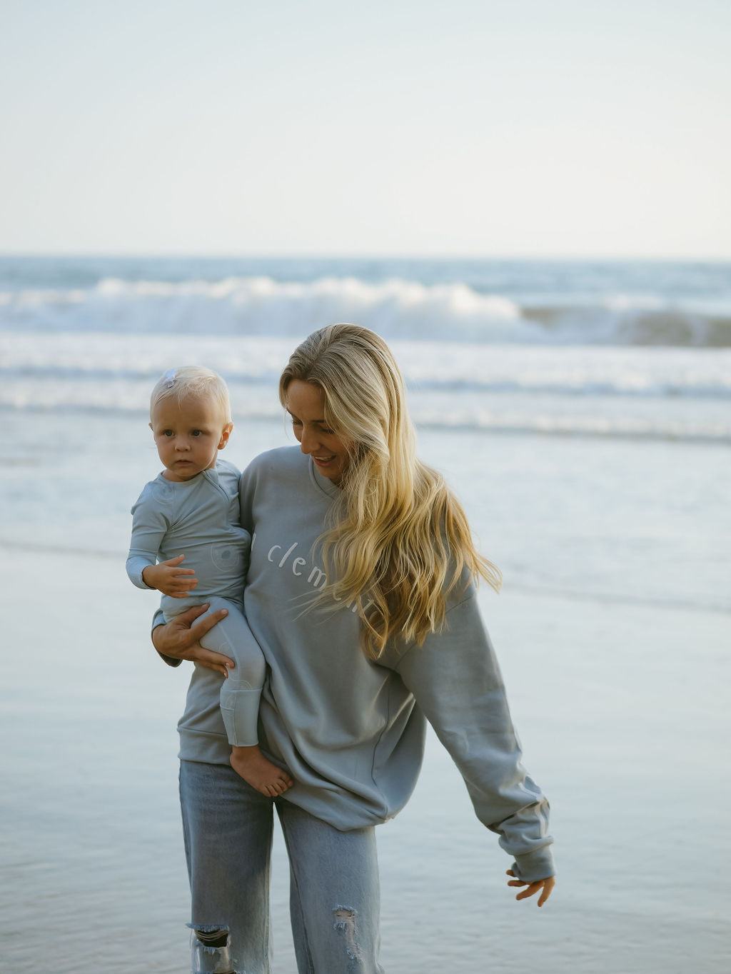 A woman with long blonde hair wears the forever french baby Adult Sweatshirt | San Clemente in light blue, holding a young child in matching clothing at the beach on wet sand with gentle waves and the ocean behind them.
