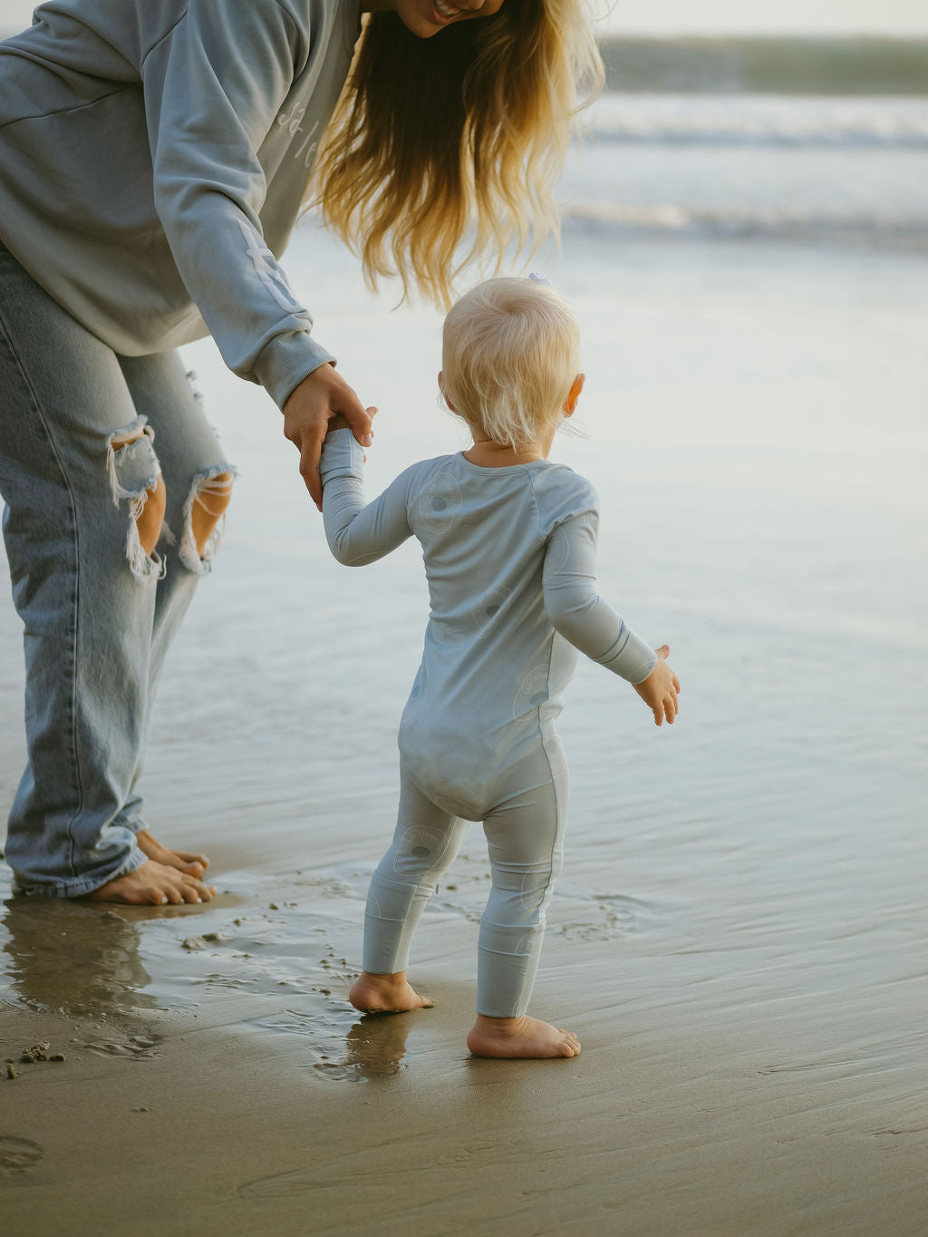 An adult holds hands with a toddler dressed in forever french baby Bamboo Zip Pajamas | San Clemente as they stand barefoot on wet beach sand, gentle waves in the background.