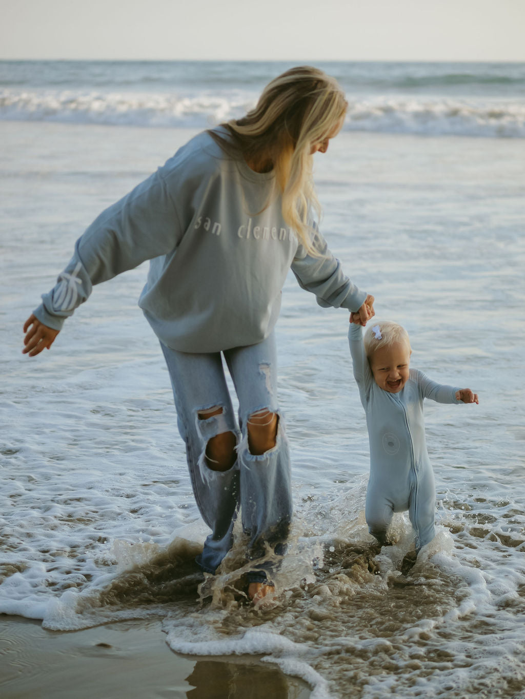 A woman in a blue sweatshirt and ripped jeans walks hand-in-hand with a smiling toddler dressed in forever french baby Bamboo Zip Pajamas | San Clemente as waves splash around their feet on the shore.