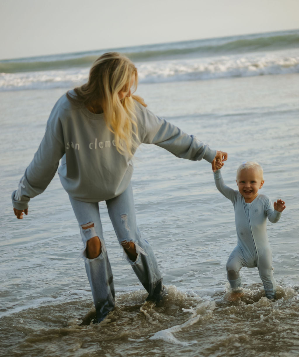 A woman wearing the forever french baby Adult Sweatshirt | San Clemente holds hands with a smiling toddler in a light blue outfit as they walk together in shallow ocean water at the beach.