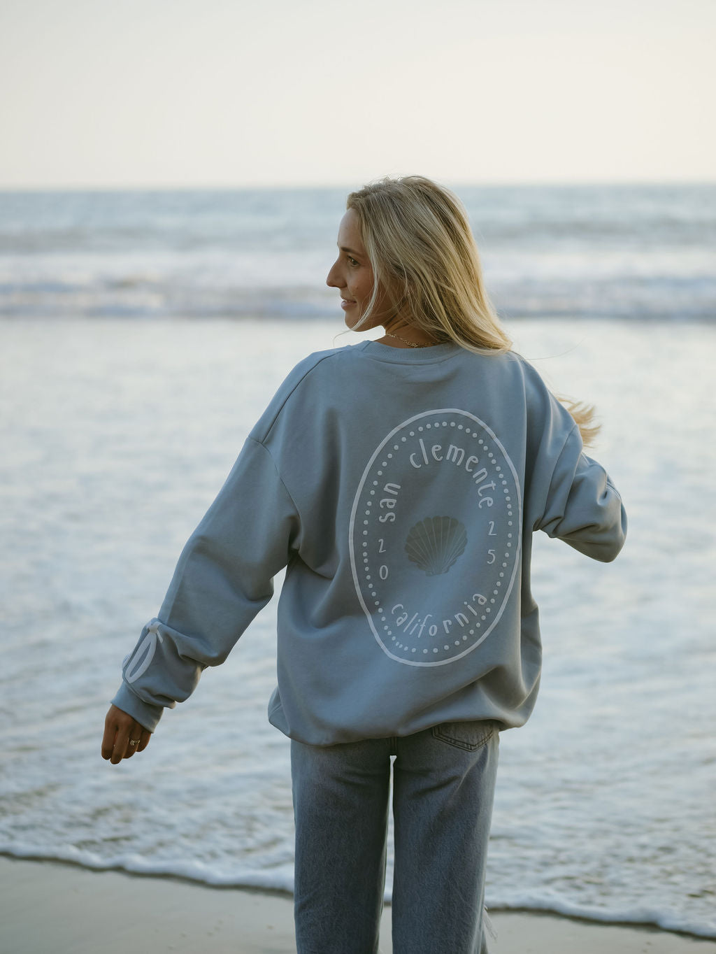 A woman with long blonde hair stands on a beach facing the ocean, wearing a light blue forever french baby Adult Sweatshirt | San Clemente with a circular seashell design on the back and light blue jeans.