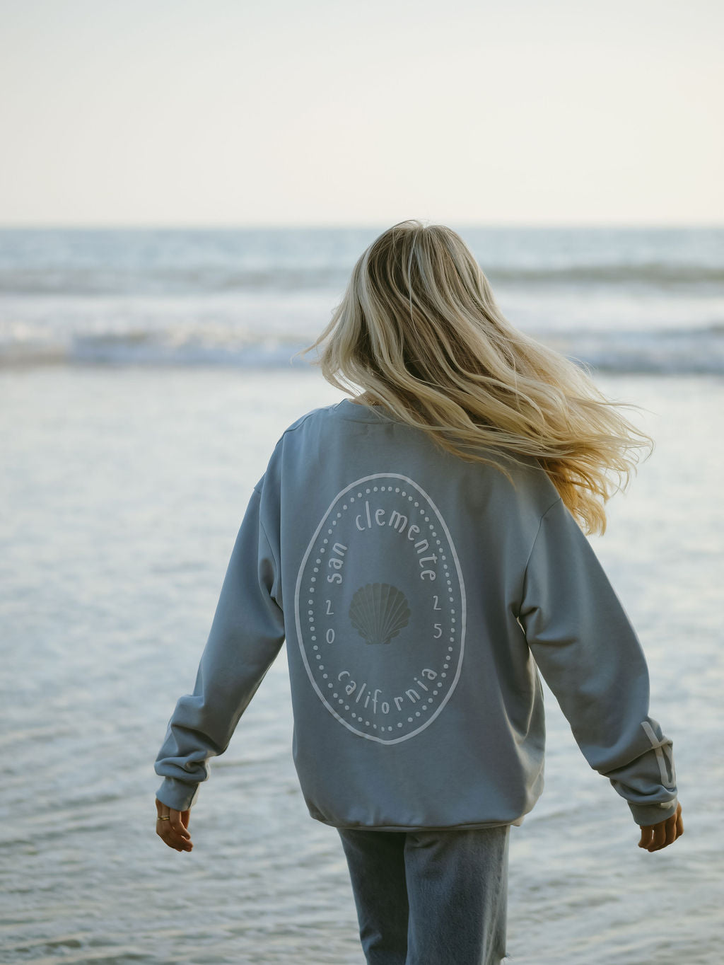 A person with long blonde hair wearing the forever french baby Adult Sweatshirt | San Clemente stands on the shore, gazing at the ocean with gentle waves and a clear sky in the background.