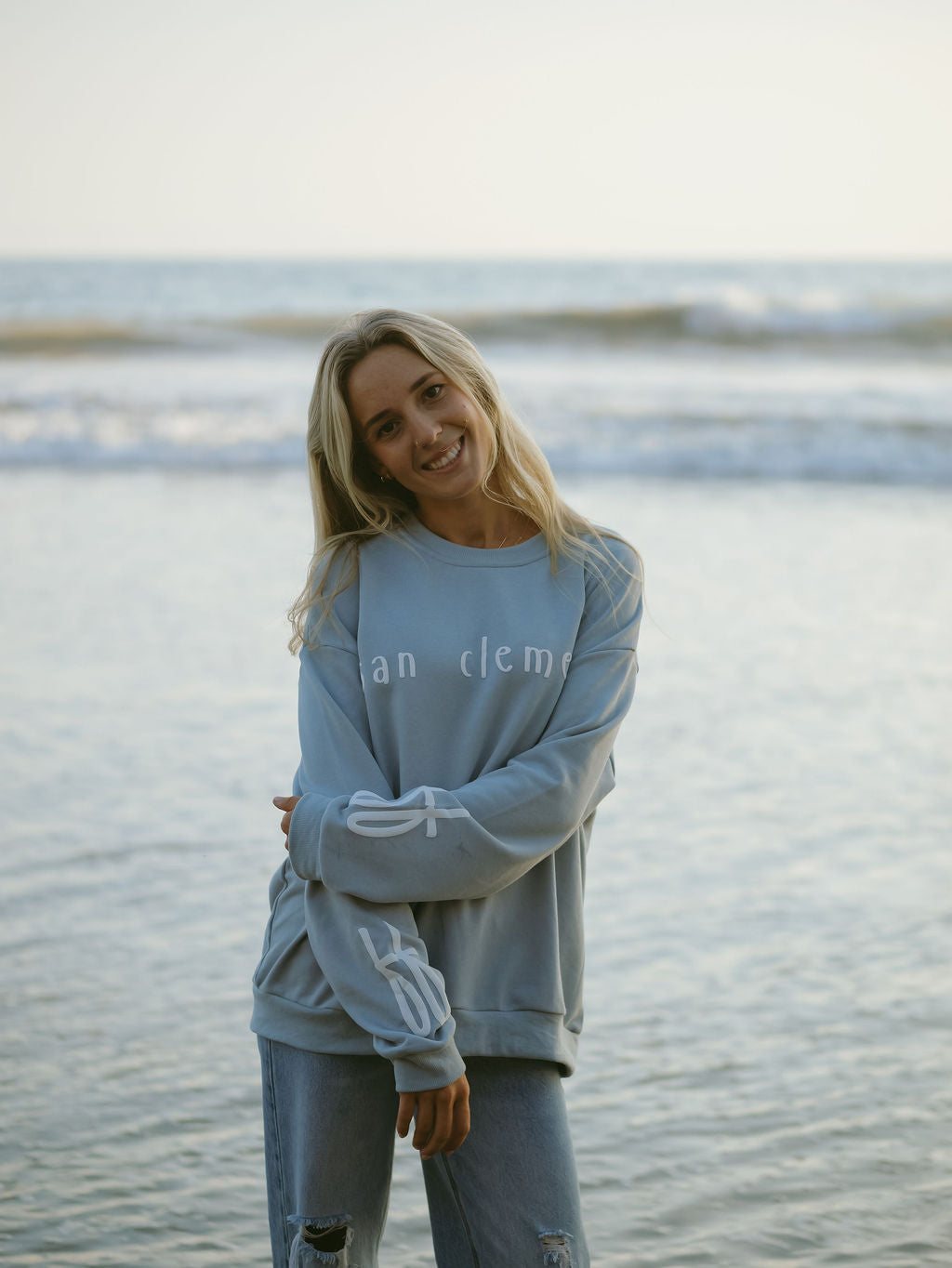 A smiling woman with long blonde hair stands by the ocean, wearing the forever french baby Adult Sweatshirt | San Clemente in light blue and ripped jeans. Gentle waves and water are visible in the background.