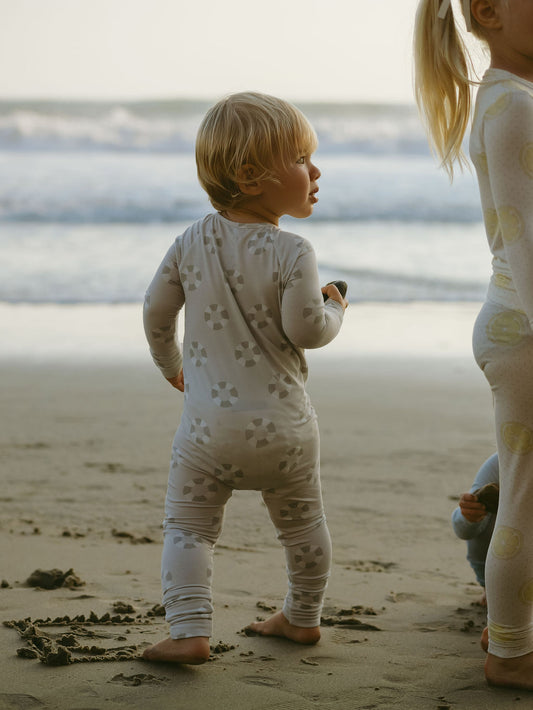 A young child wears forever french baby Bamboo Zip Pajamas | Lifesaver with geometric patterns, standing barefoot on a sandy beach and gazing at the ocean. Another child stands nearby as gentle waves roll in under soft light.