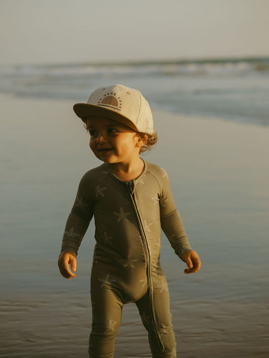 A toddler in forever french baby's Bamboo Zip Pajamas | Starfish and a beige cap stands on the beach, smiling and looking to the side with waves in the background.