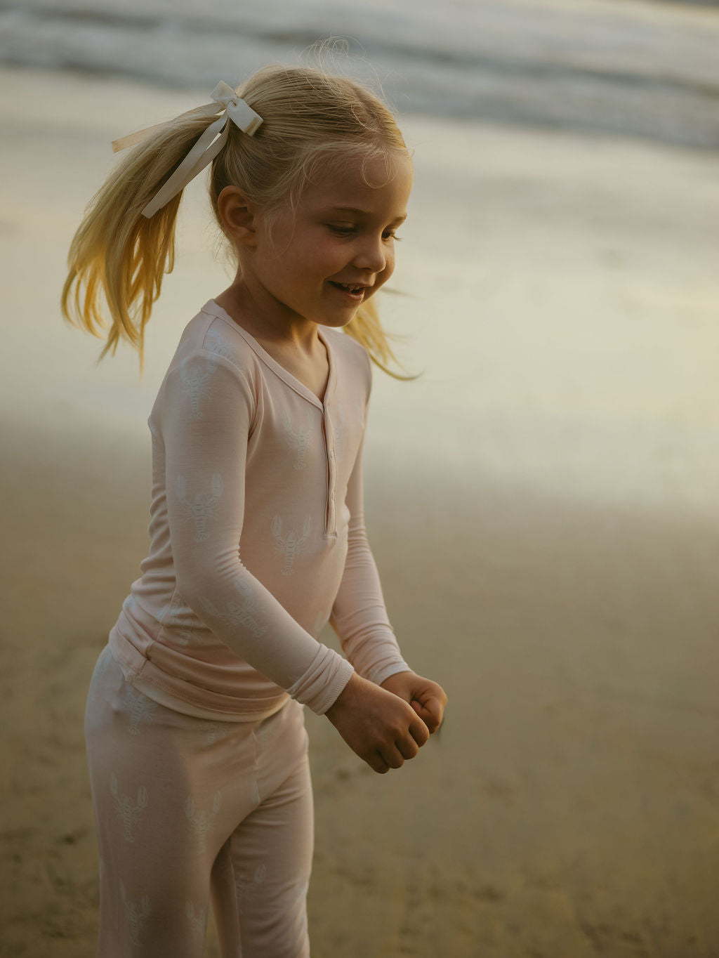 A young blonde girl in pigtails wears forever french baby's Bamboo Two Piece Pajamas | Lobster—light pink, hypoallergenic kids sleepwear with a subtle pattern—while smiling and playing on the beach at sunset.