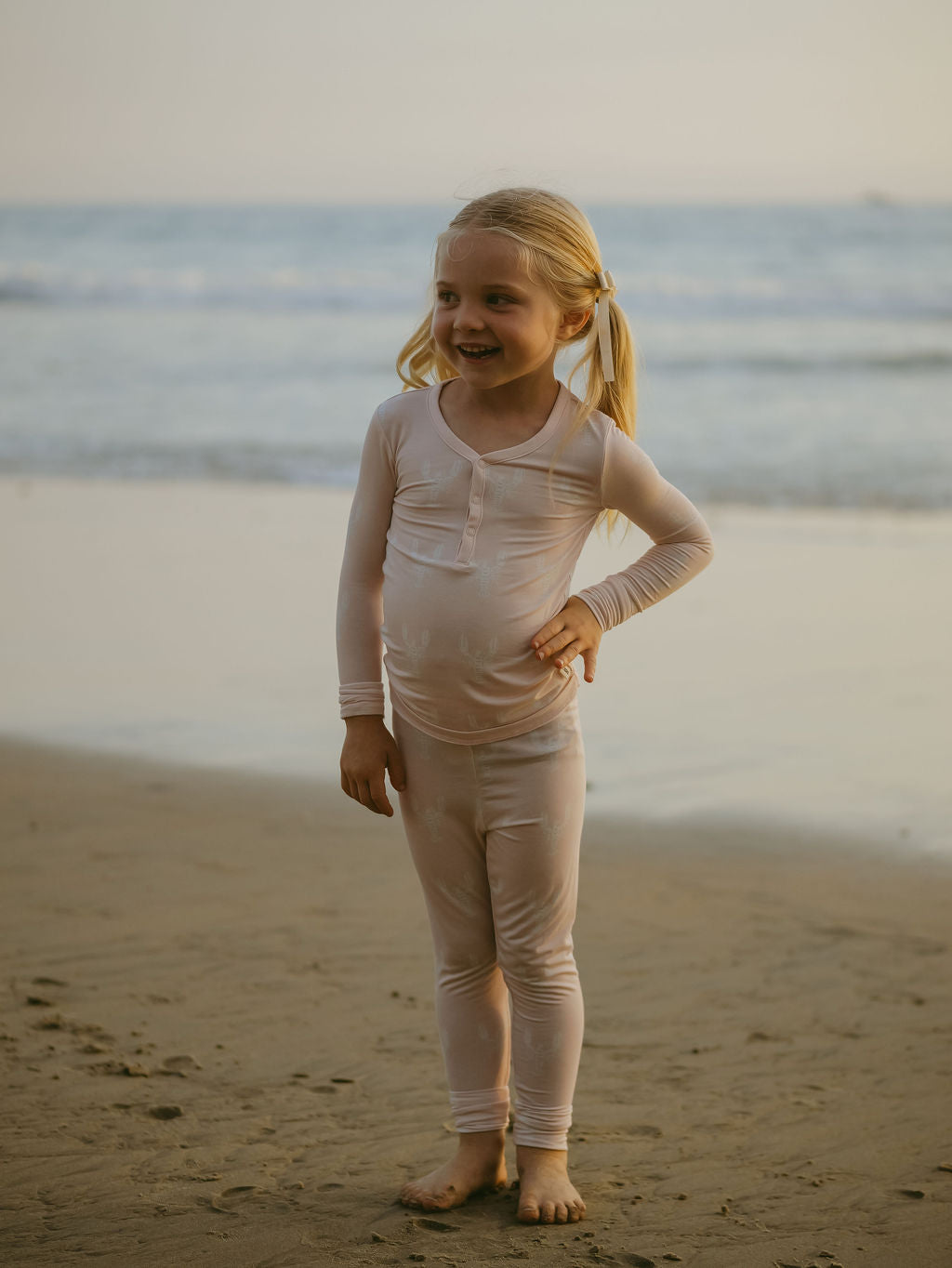 A smiling blonde girl in pigtails poses barefoot on the beach, ocean behind her, wearing forever french baby Bamboo Two Piece Pajamas | Lobster—breathable, comfy children's sleepwear.