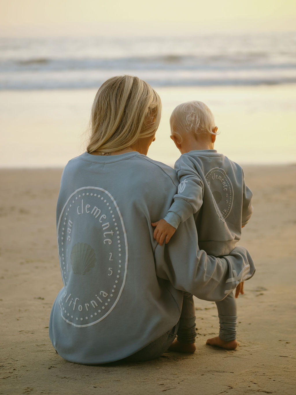 A woman and toddler with blonde hair sit on the beach at sunset, both wearing forever french baby's Child Sweatshirt | San Clemente—matching blue kids clothing with a seashell graphic and “San Clemente, California” on the back.