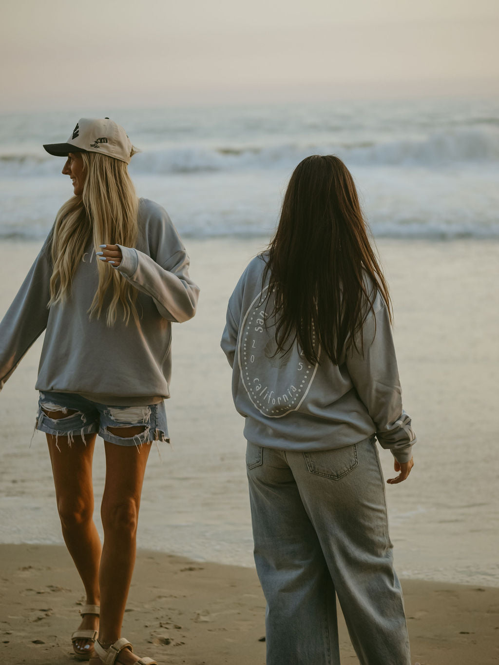 Two women in casual wear—one in a cap and shorts, the other in jeans and a forever french baby San Clemente Adult Sweatshirt—stand on a sandy beach at sunset, gazing at the ocean as waves roll in.