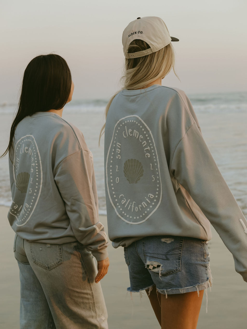 Two women stand on the beach in matching forever french baby Adult Sweatshirt | San Clemente tops with a shell design. Paired with denim, their look and the pastel sky capture a relaxed California coastal vibe.