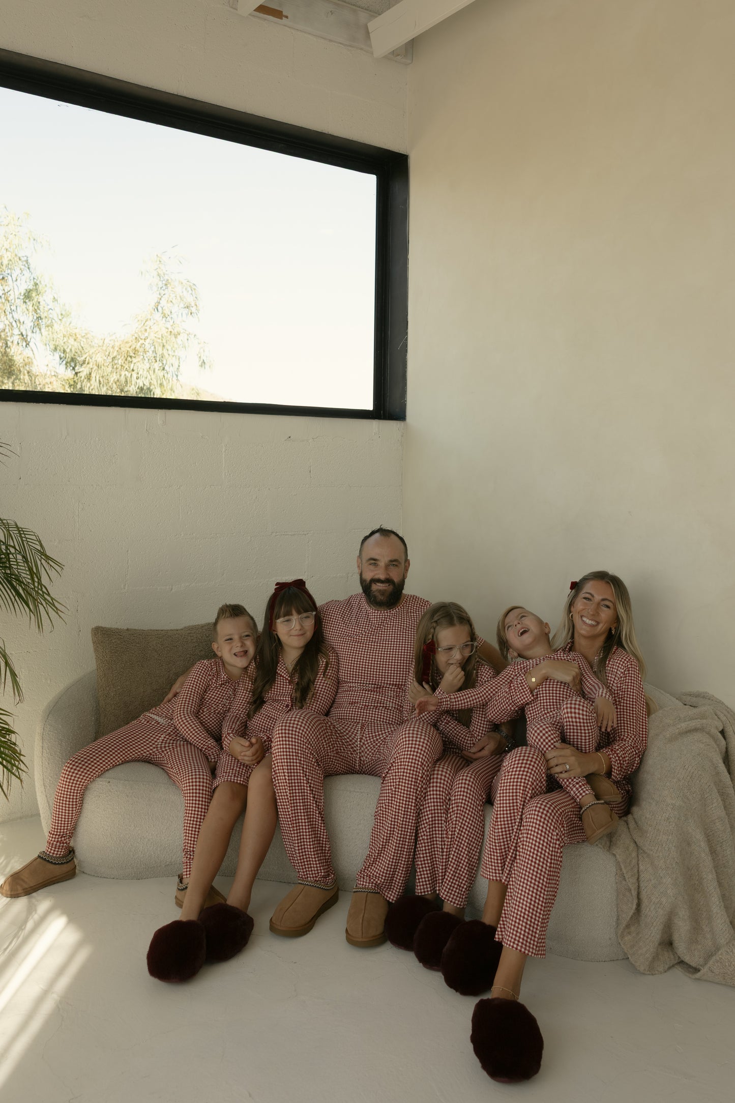 A family of five cozies up on a couch, all in forever french baby’s Men’s Short Sleeve Bamboo Pajamas in red gingham, smiling and cuddling together in their modern living room.