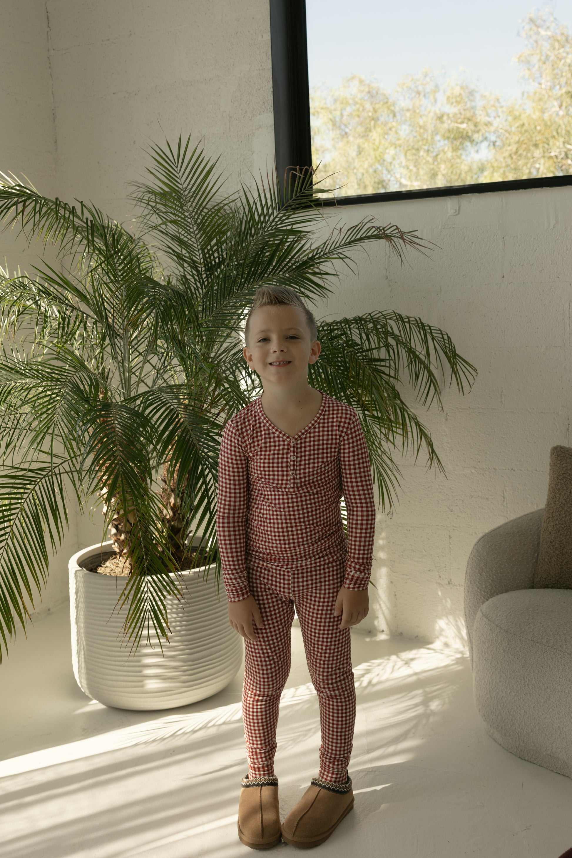 A young child smiles indoors in sunlight, wearing forever french baby’s Bamboo Two Piece Pajamas in Red Gingham and brown slippers, standing by a large potted palm.