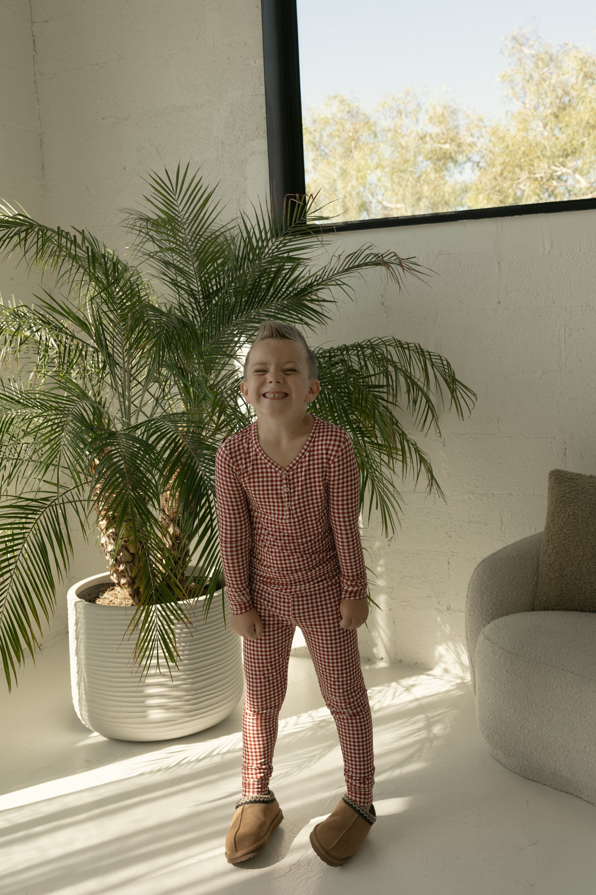 A child in forever french baby Bamboo Two Piece Pajamas | Red Gingham stands indoors by a large potted palm, sunlight streaming through the window and brightening the modern room.