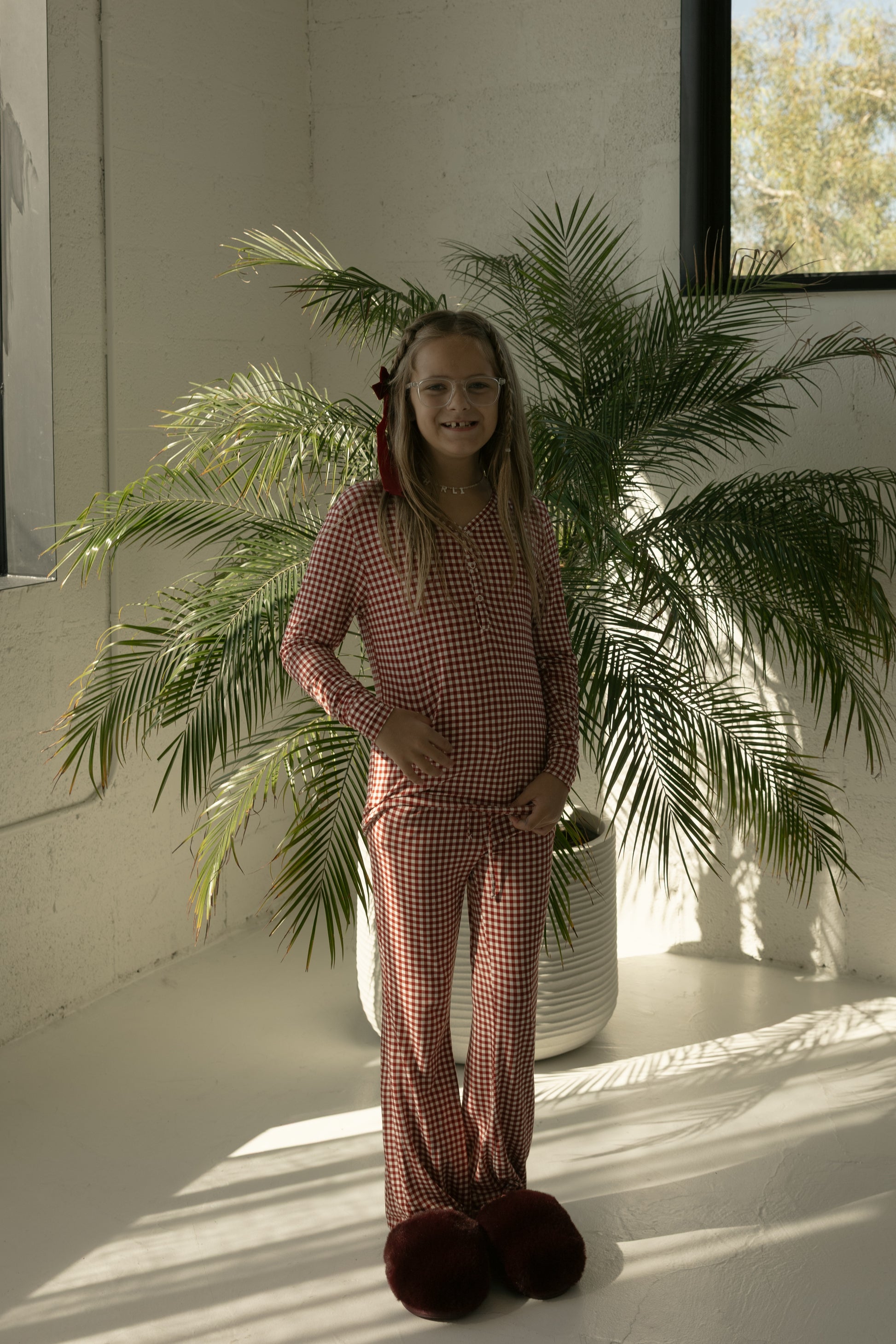 A smiling pre-teen girl with long hair and glasses wears forever french baby’s Pre-Teen Flare Bamboo Pajamas in Red Gingham, standing indoors by a large potted palm as sunlight shines through the window.