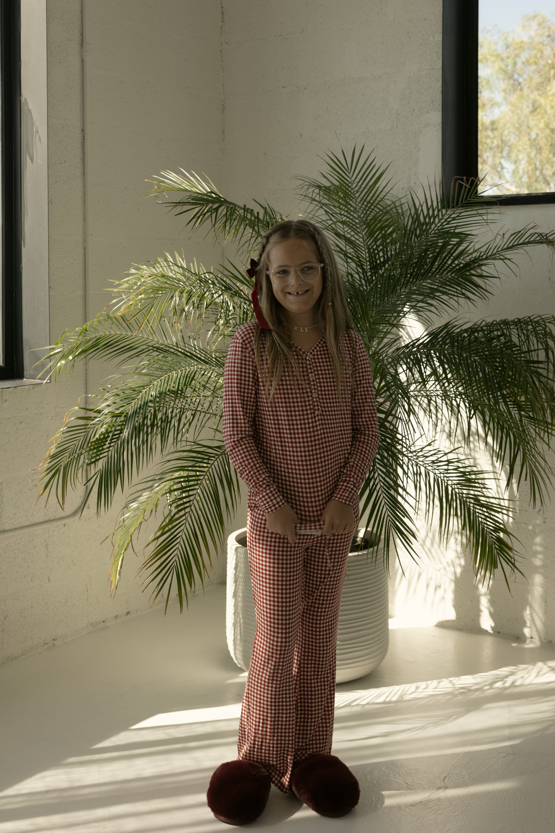 A smiling young girl poses in forever french baby’s Pre-Teen Flare Bamboo Pajamas in Red Gingham, paired with fuzzy slippers, standing by a potted palm in a bright room with white walls and large windows.