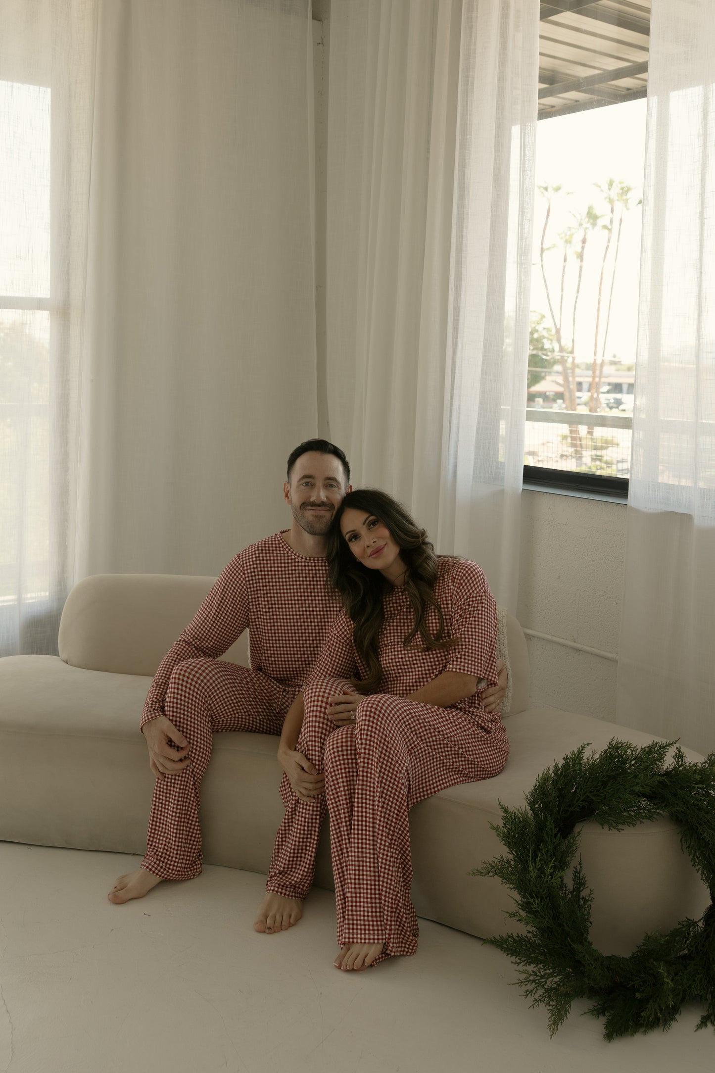 A couple in forever french baby Men's Bamboo Pajamas | Red Gingham sits on a beige couch in a bright room with white curtains, the woman leaning on the man's shoulder and a green wreath resting on the floor beside them.