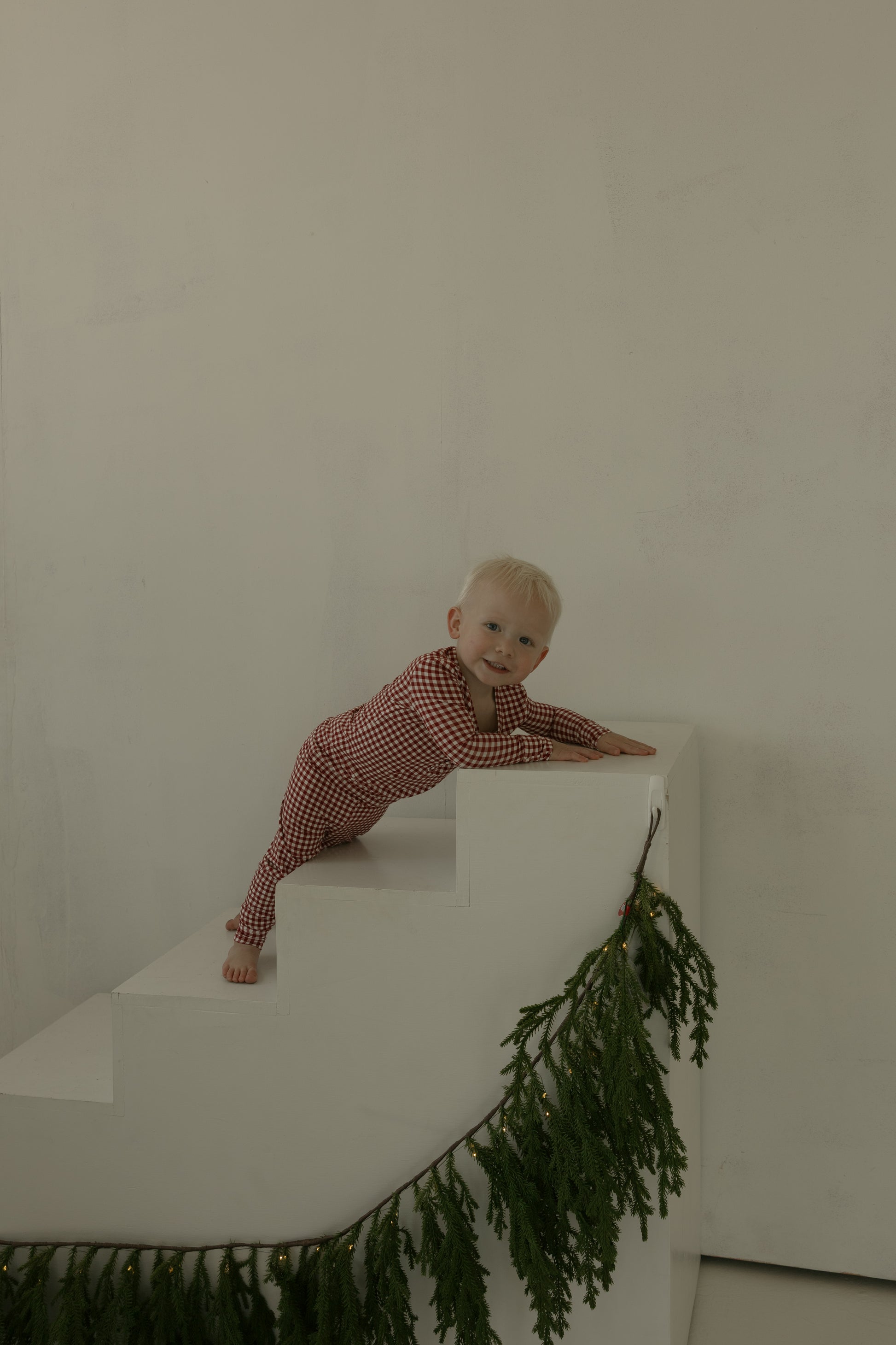 A baby in forever french baby's Bamboo Two Piece Pajamas in Red Gingham crawls up white stairs with green pine garland, against a plain light wall—adorable, comfy holiday kids' sleepwear.