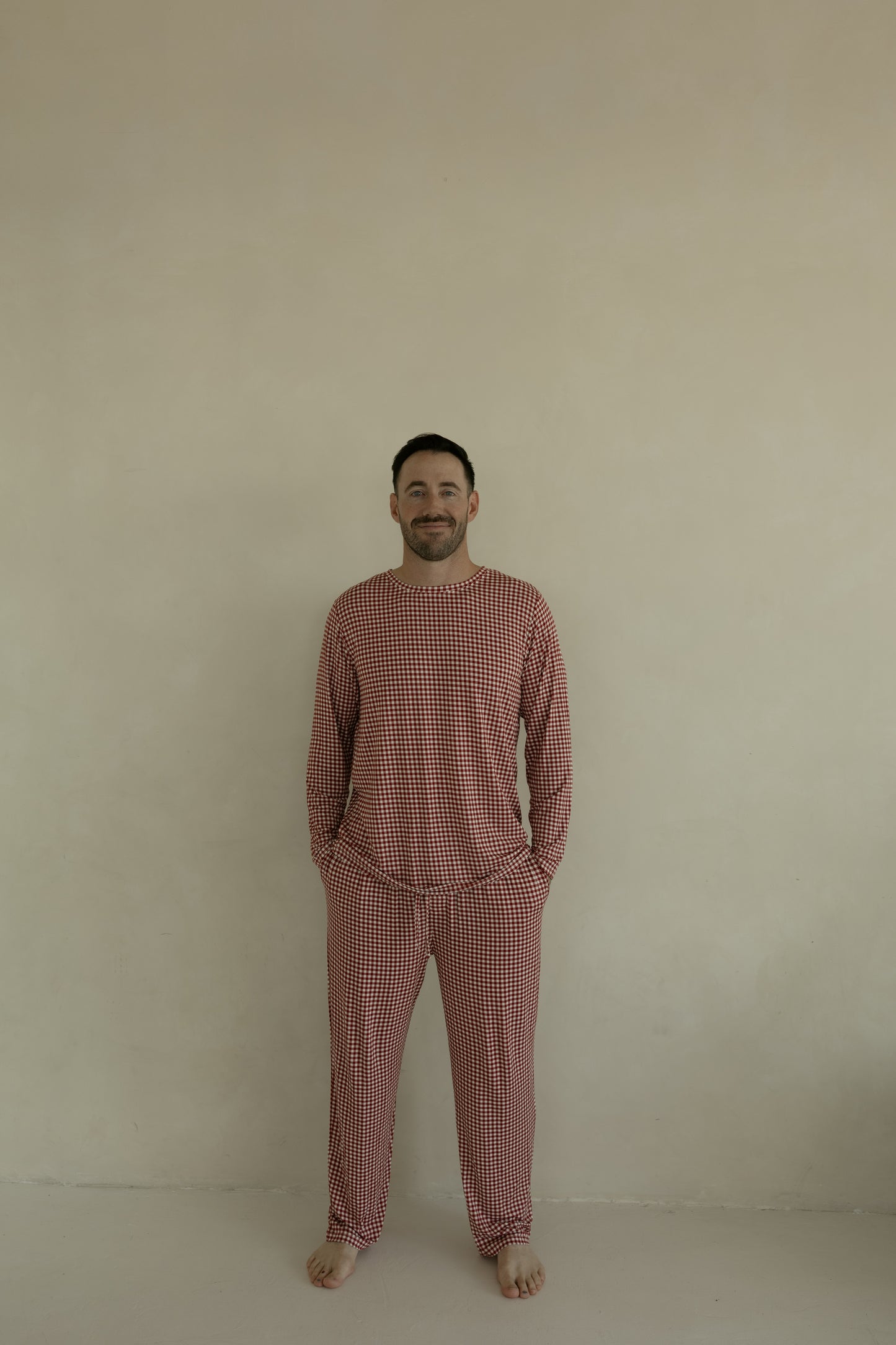 A man stands barefoot against a beige wall, smiling as he wears forever french baby Men's Bamboo Pajamas in Red Gingham. His hands in his pockets, he enjoys the hypo-allergenic comfort of bamboo sleepwear.