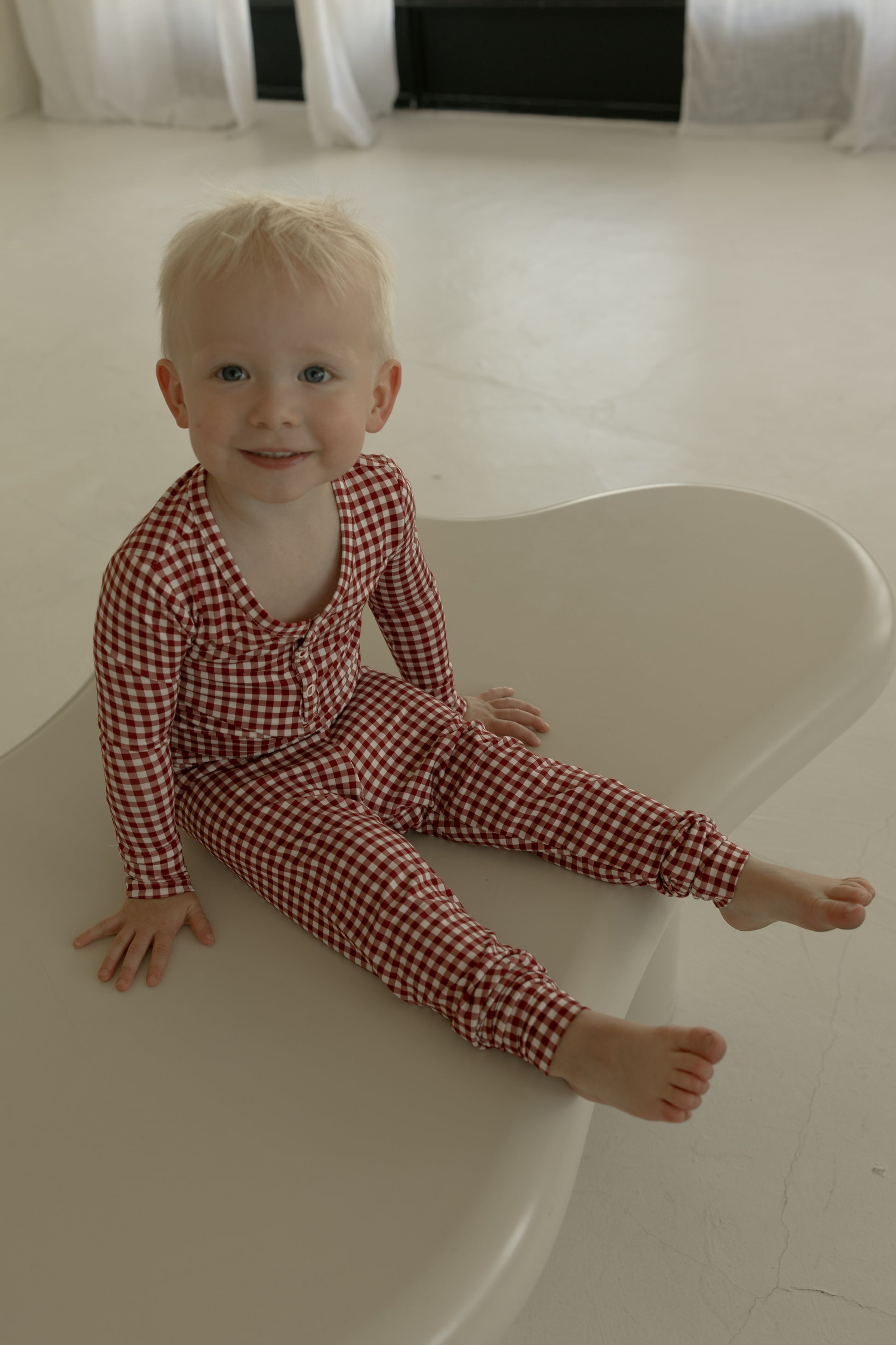 A young child with blonde hair, barefoot, wears forever french baby’s Bamboo Two Piece Pajamas in Red Gingham while sitting on a light-colored furniture piece in a bright, minimal room.