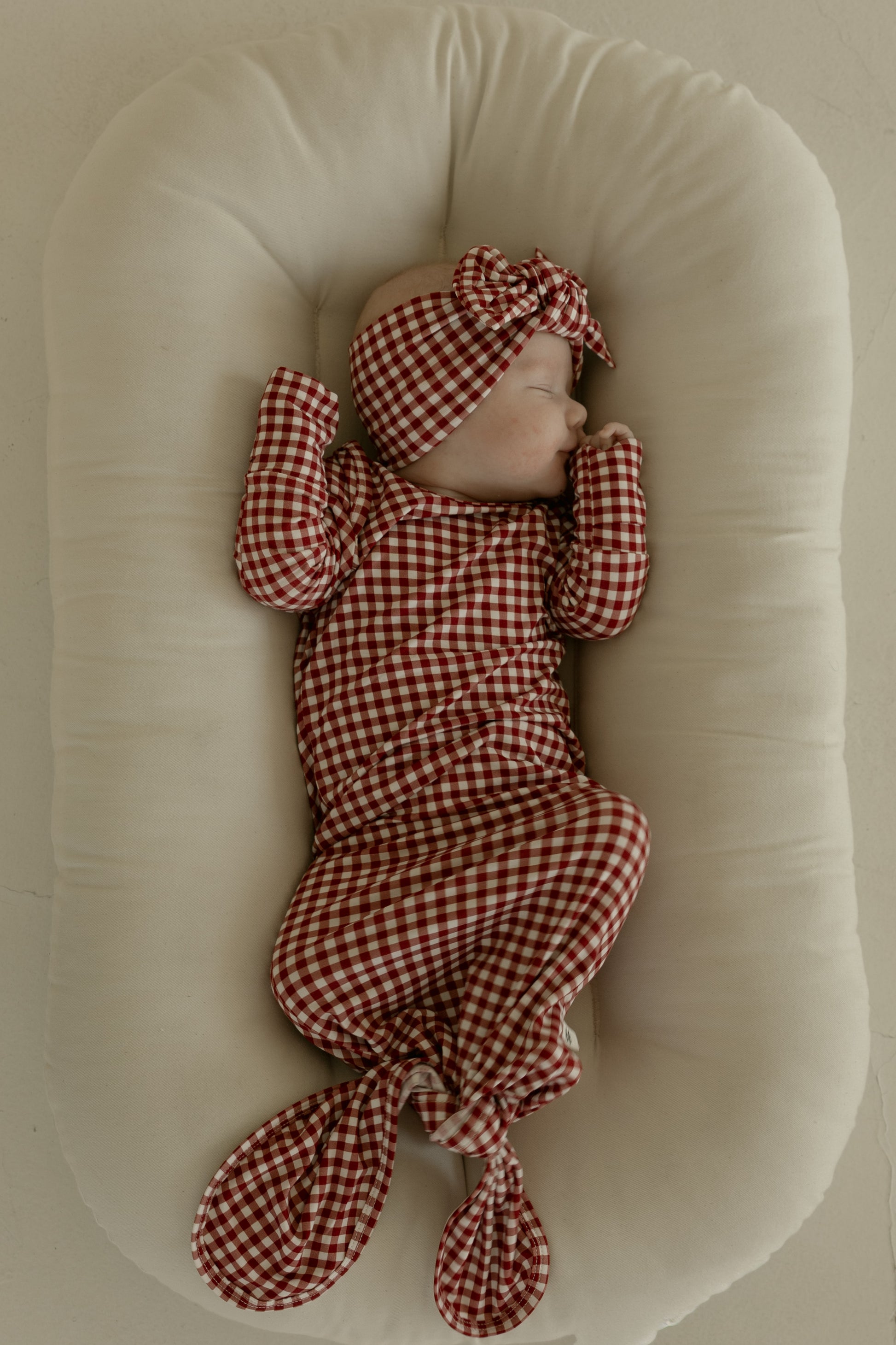 A baby sleeps on a soft, cream-colored cushion, wearing the forever french baby Bamboo Knotted Gown in Red Gingham, lying on their back with matching hat and footed pants, arms bent and eyes closed.
