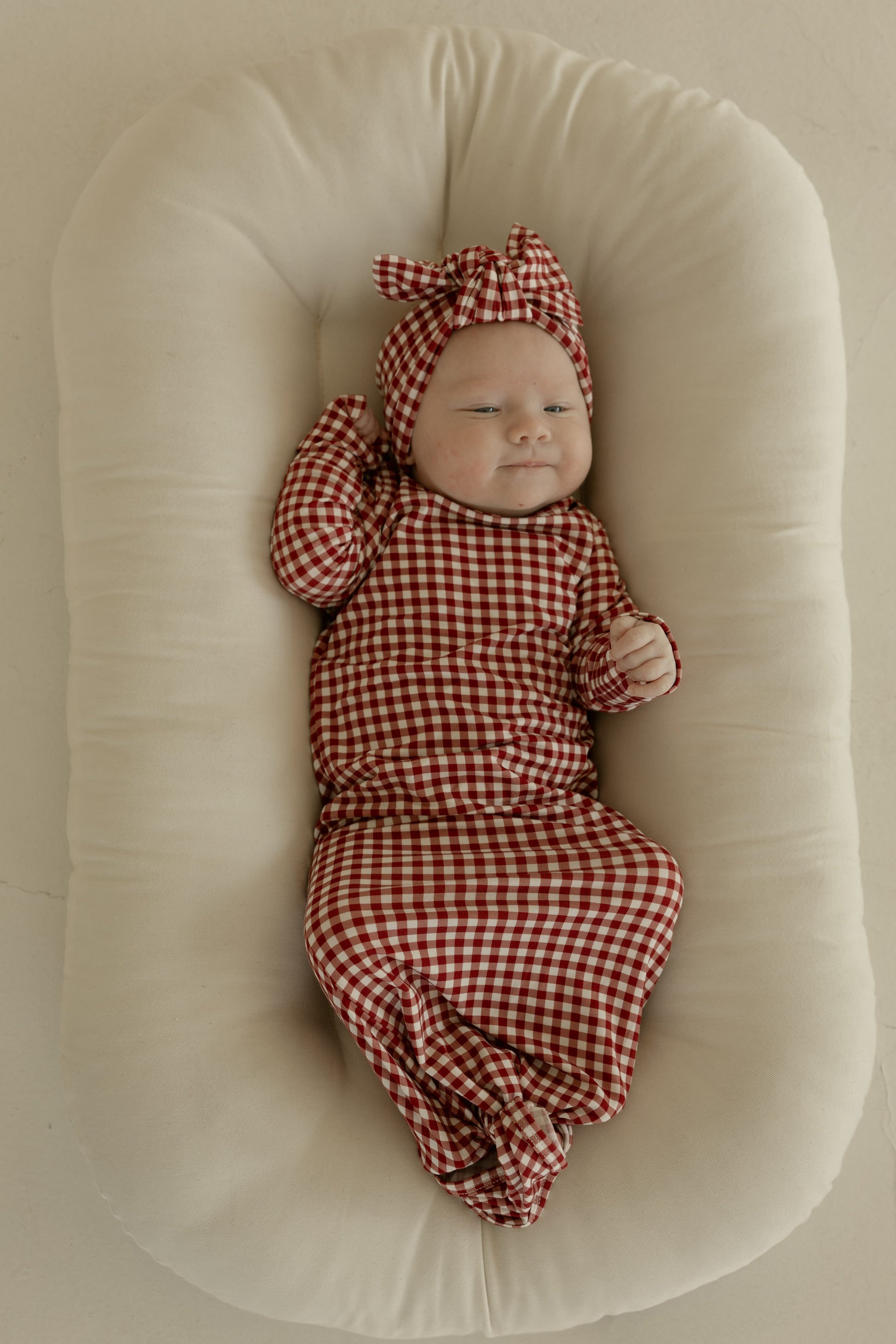 A baby smiles softly on a cream pillow, wearing the forever french baby Bamboo Knotted Gown in Red Gingham with a matching headband and bow, one arm raised in comfort.