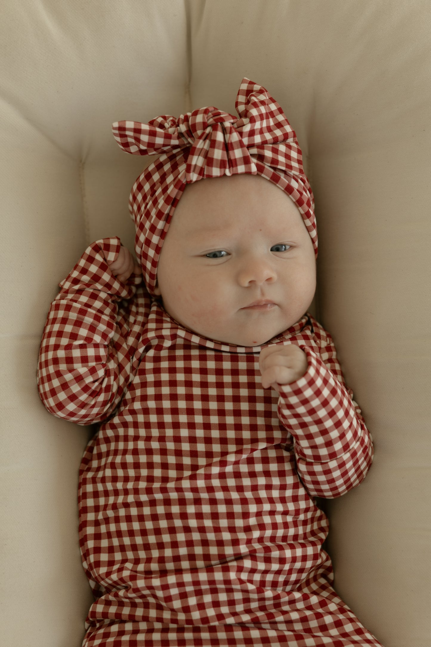 A baby lies on a light cushion, wearing the forever french baby Bamboo Head Wrap in Red Gingham, paired with a matching outfit, gazing upward with a calm expression.