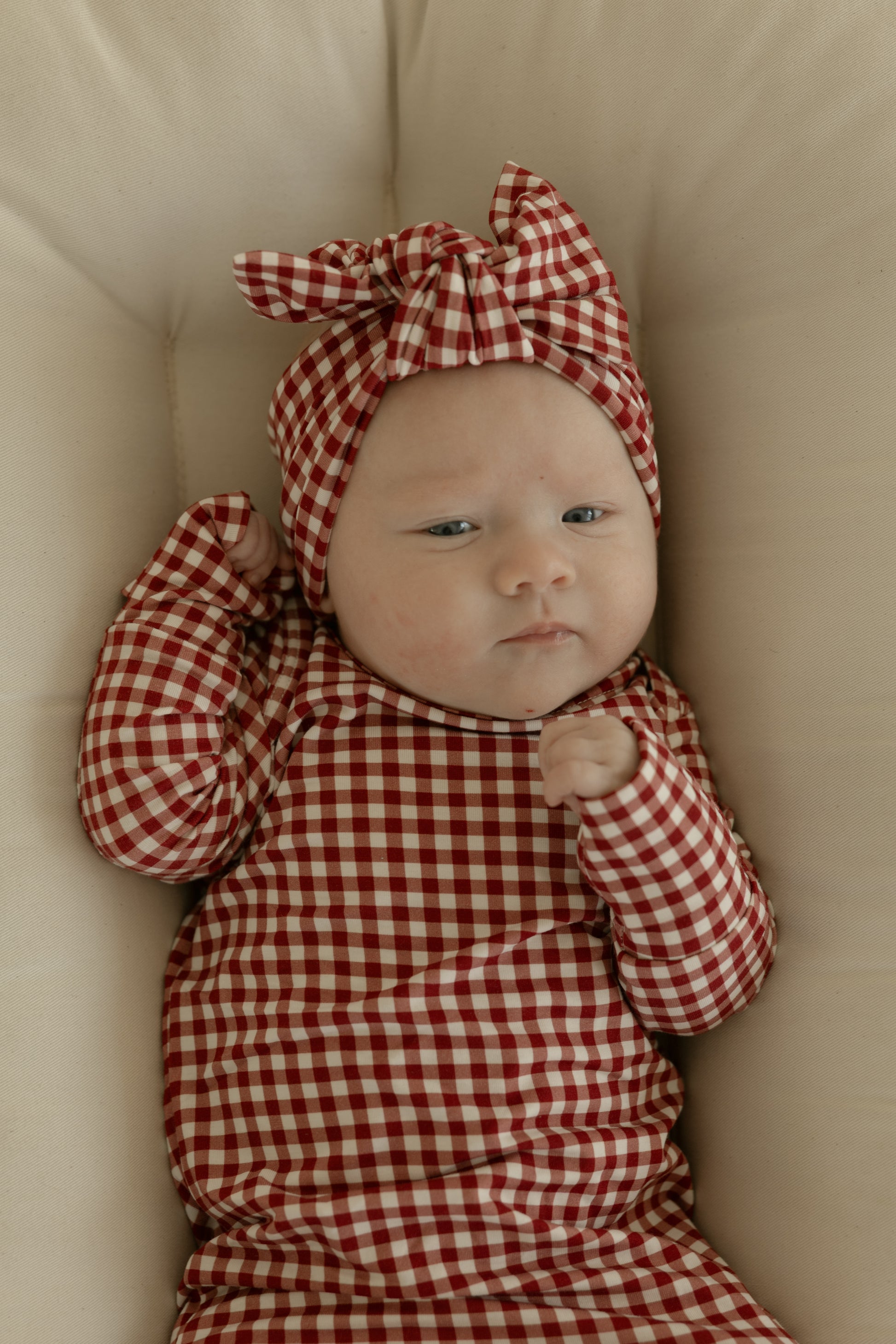 A baby lies on a light cushion, wearing the forever french baby Bamboo Head Wrap in Red Gingham, paired with a matching outfit, gazing upward with a calm expression.