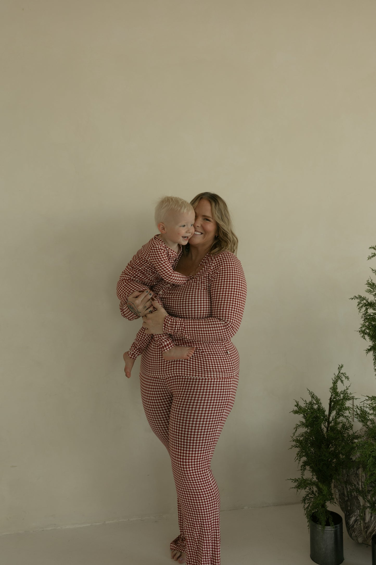 A woman and a young child, both wearing matching forever french baby Women's Bamboo Pajamas in Red Gingham, smile together in front of a beige wall with a green potted plant nearby.