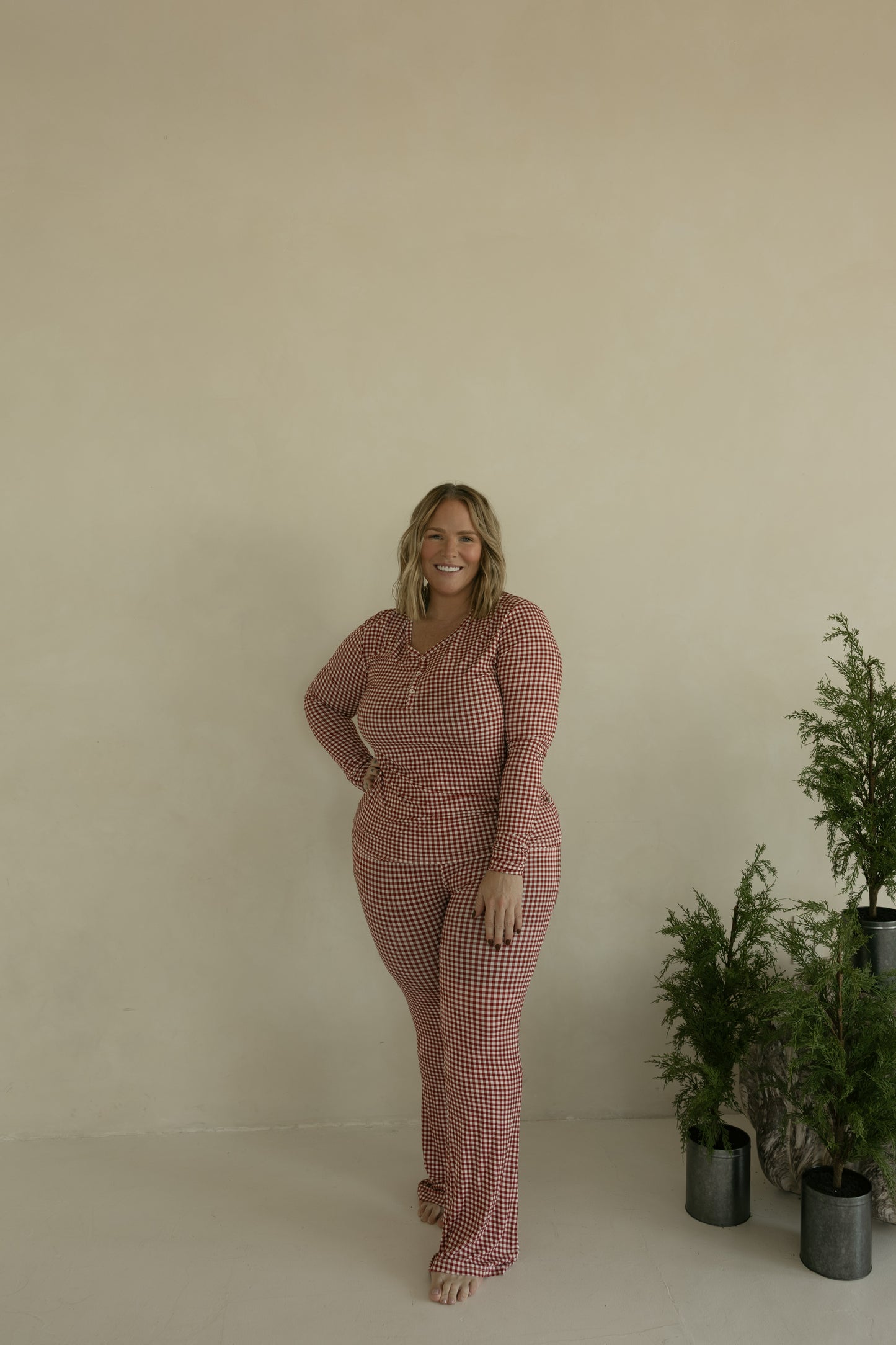 A woman wears forever french baby’s Women’s Bamboo Pajamas in Red Gingham, standing barefoot in a minimal beige room with small green potted plants to her right. She smiles confidently, hand on her hip. Pajamas are breastfeeding friendly.