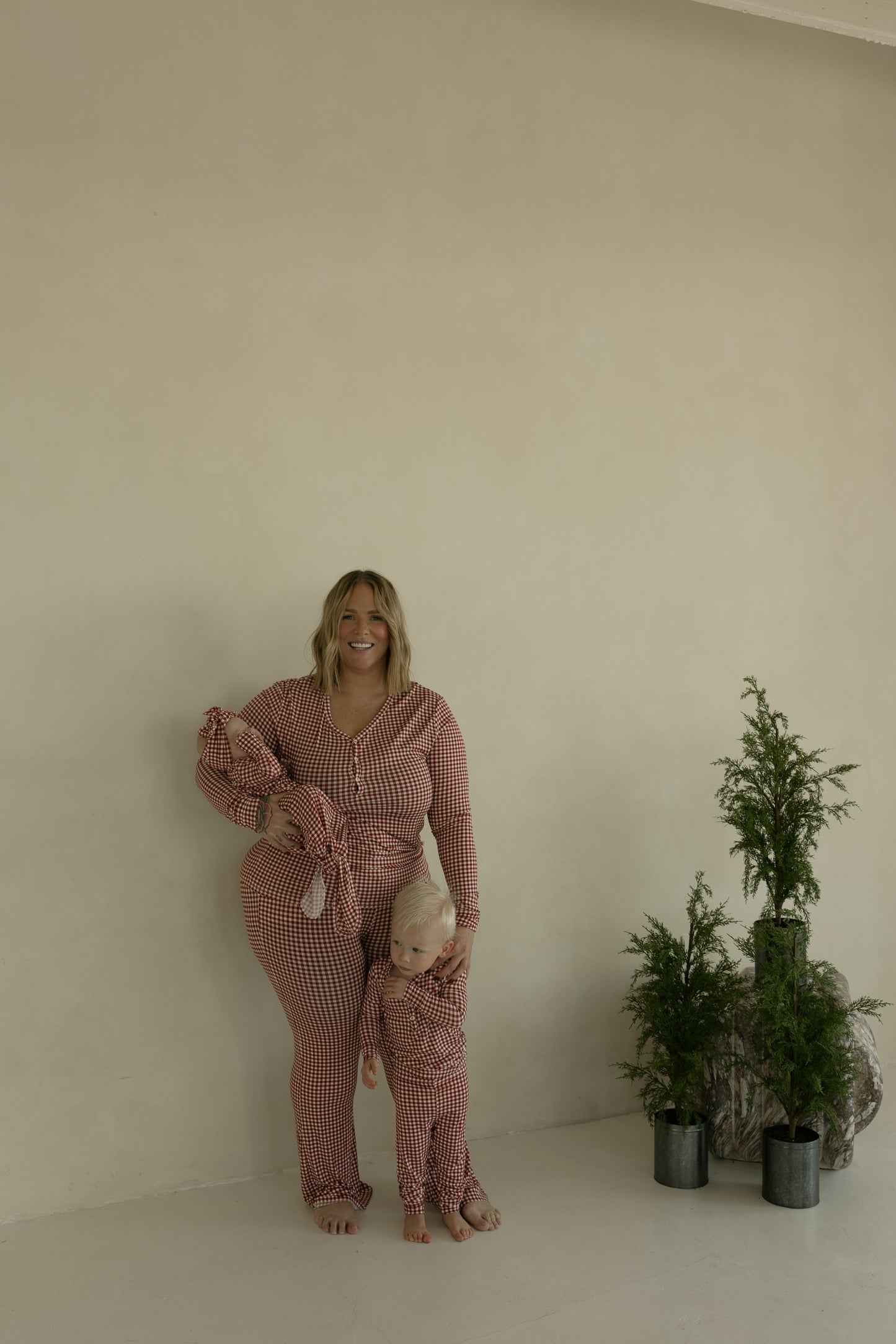 A woman and child, barefoot, wear matching forever french baby Women's Bamboo Pajamas in Red Gingham as they stand by two potted plants against a beige wall. The woman smiles warmly, holding the child close.