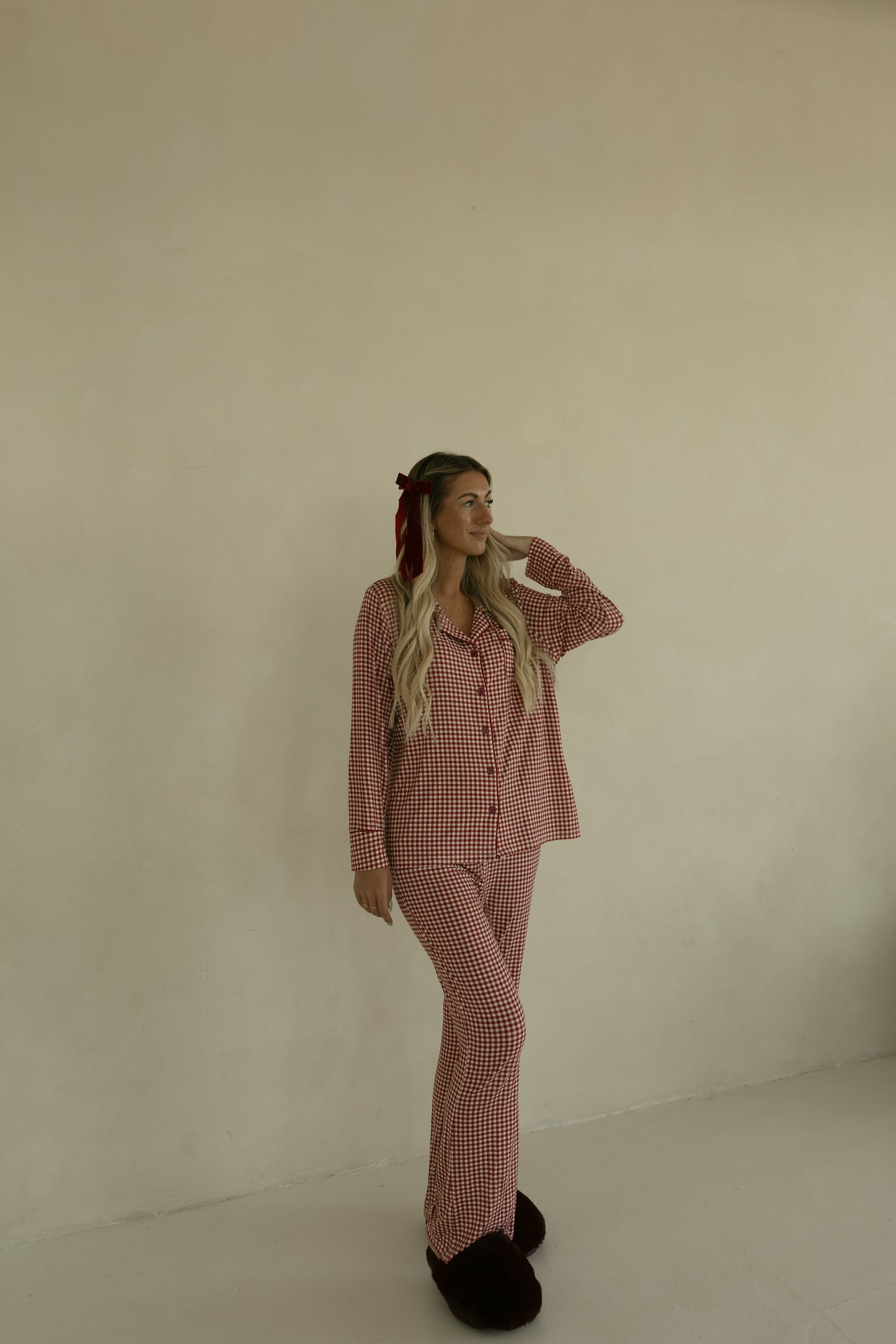 A woman with long blonde hair, wearing forever french baby’s Long Sleeve Women's Bamboo Pajamas in Red Gingham, dark slippers, and a red hair ribbon stands against a plain beige wall, looking to her left with a relaxed expression.