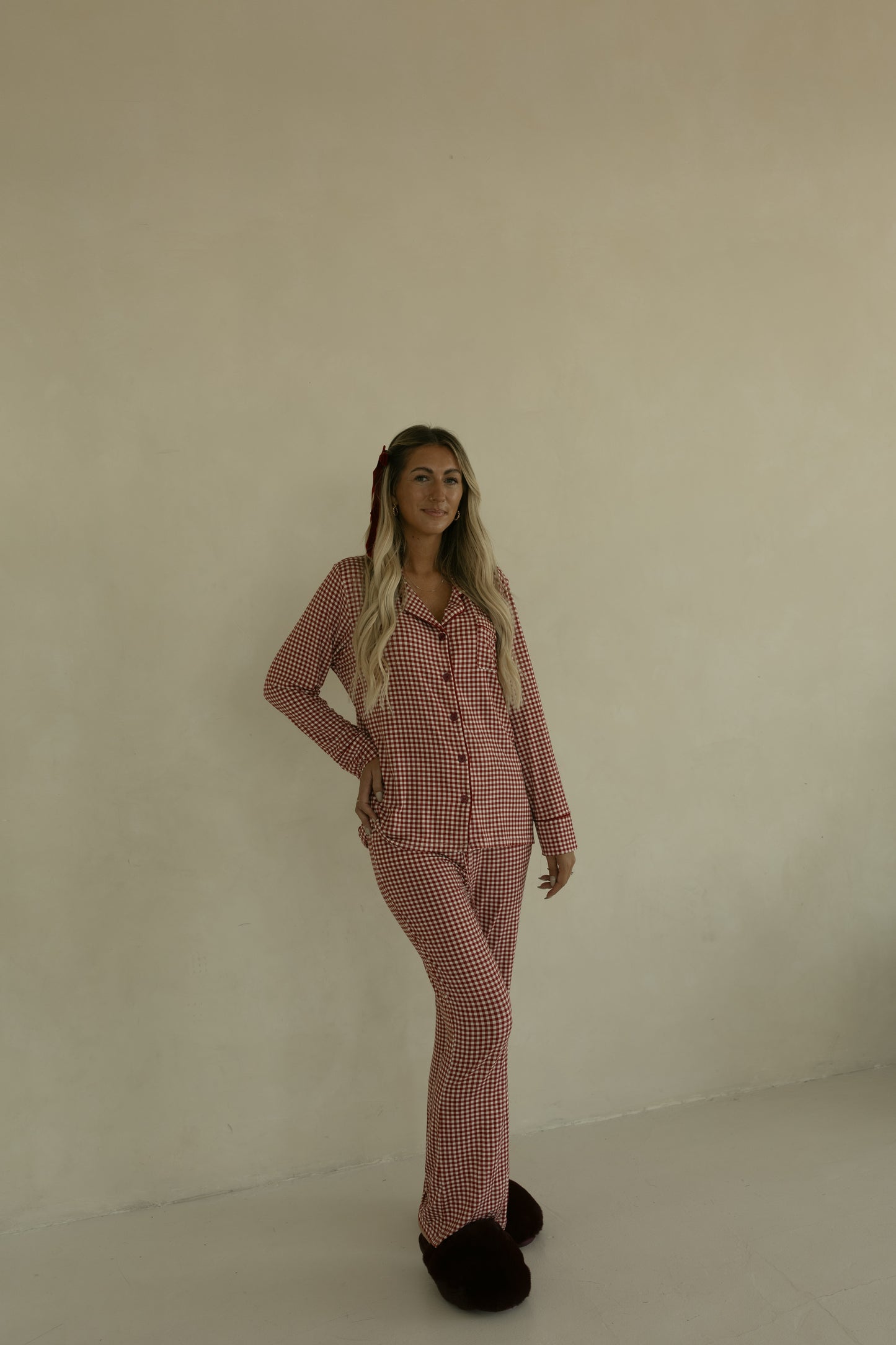 A woman with long blonde hair, smiling slightly with one hand on her hip, stands against a beige wall wearing forever french baby’s Long Sleeve Women's Bamboo Pajamas in Red Gingham and dark slippers.