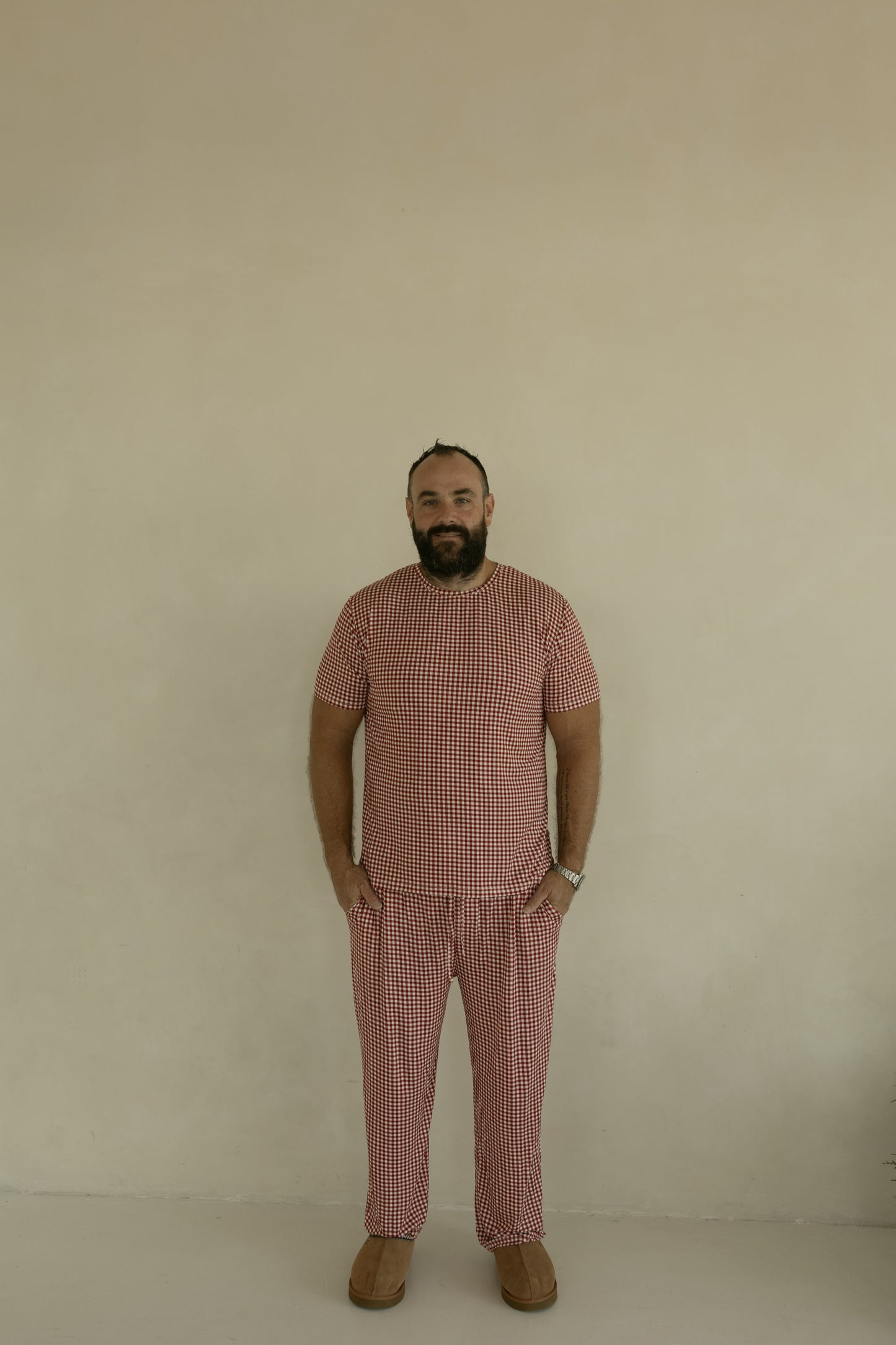 A bearded man stands against a beige wall, wearing forever french baby's Men's Short Sleeve Bamboo Pajamas in Red Gingham with brown shoes. He smiles slightly, hands in pockets, showing off these hypoallergenic pajamas.