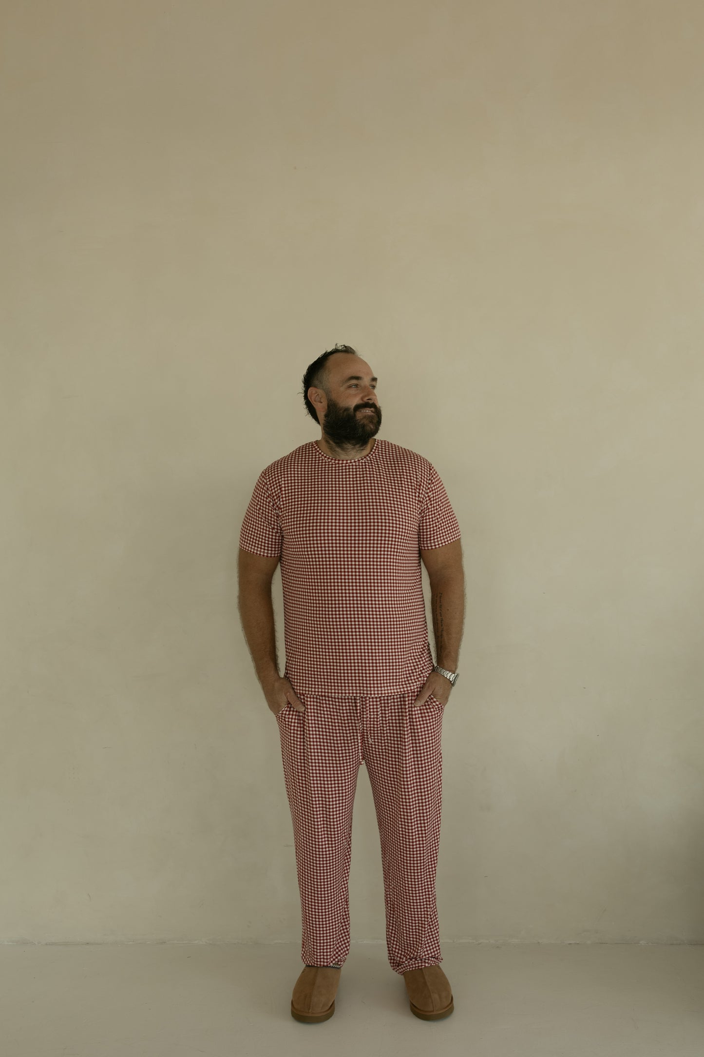 A man stands against a beige wall, wearing forever french baby's Men's Short Sleeve Bamboo Pajamas in Red Gingham. He has his hands in his pockets, looks slightly left, and pairs the set with brown shoes.