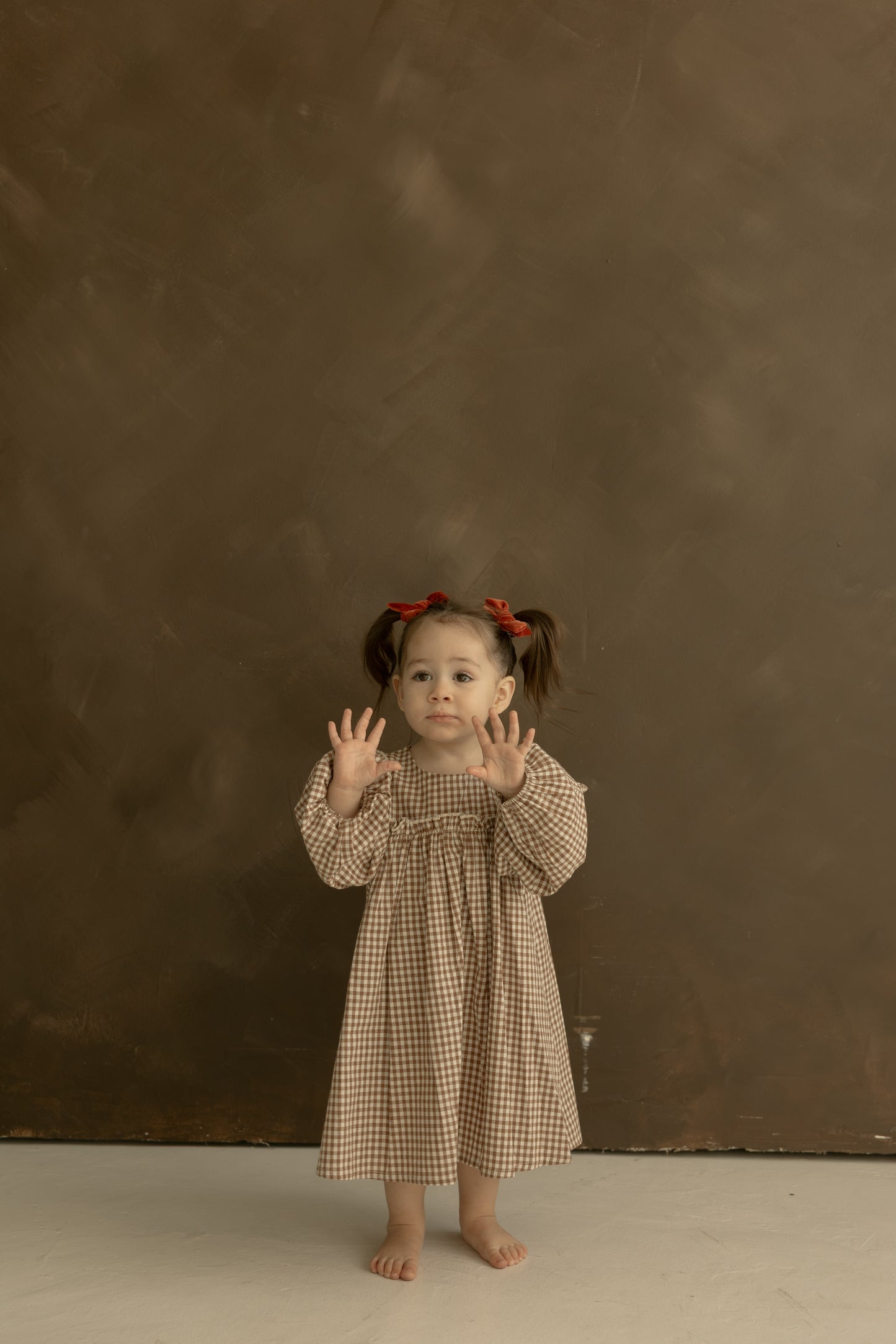 A young girl with pigtails and red bows stands barefoot on a light floor, wearing the forever french baby Child Long Sleeve Dress in Honey Gingham. She holds her hands up, fingers spread, in front of a plain brown background.