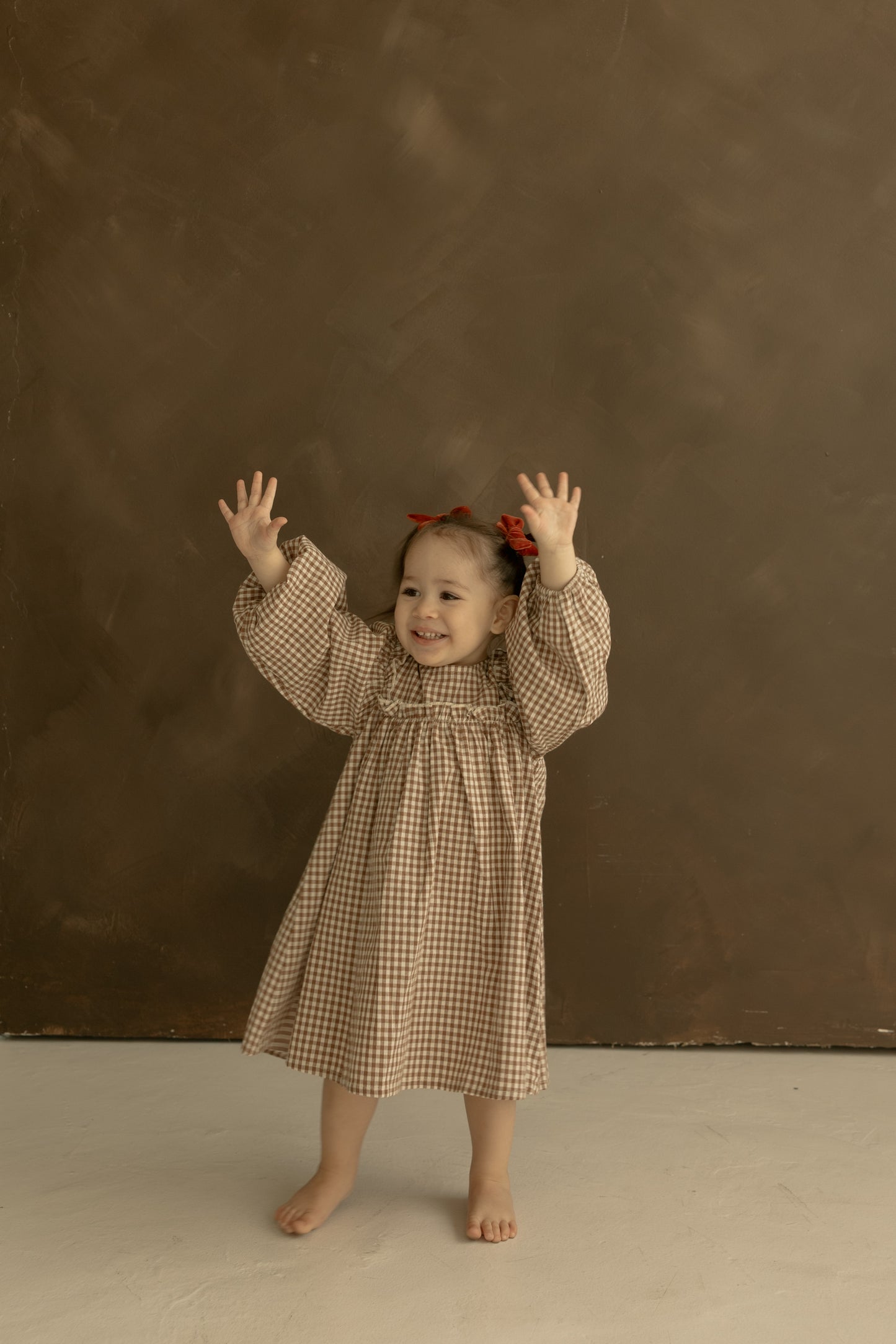 A smiling young girl wears the forever french baby Child Long Sleeve Dress in Honey Gingham, barefoot on a light floor with a red hair bow, playfully raising both hands against a brown background.