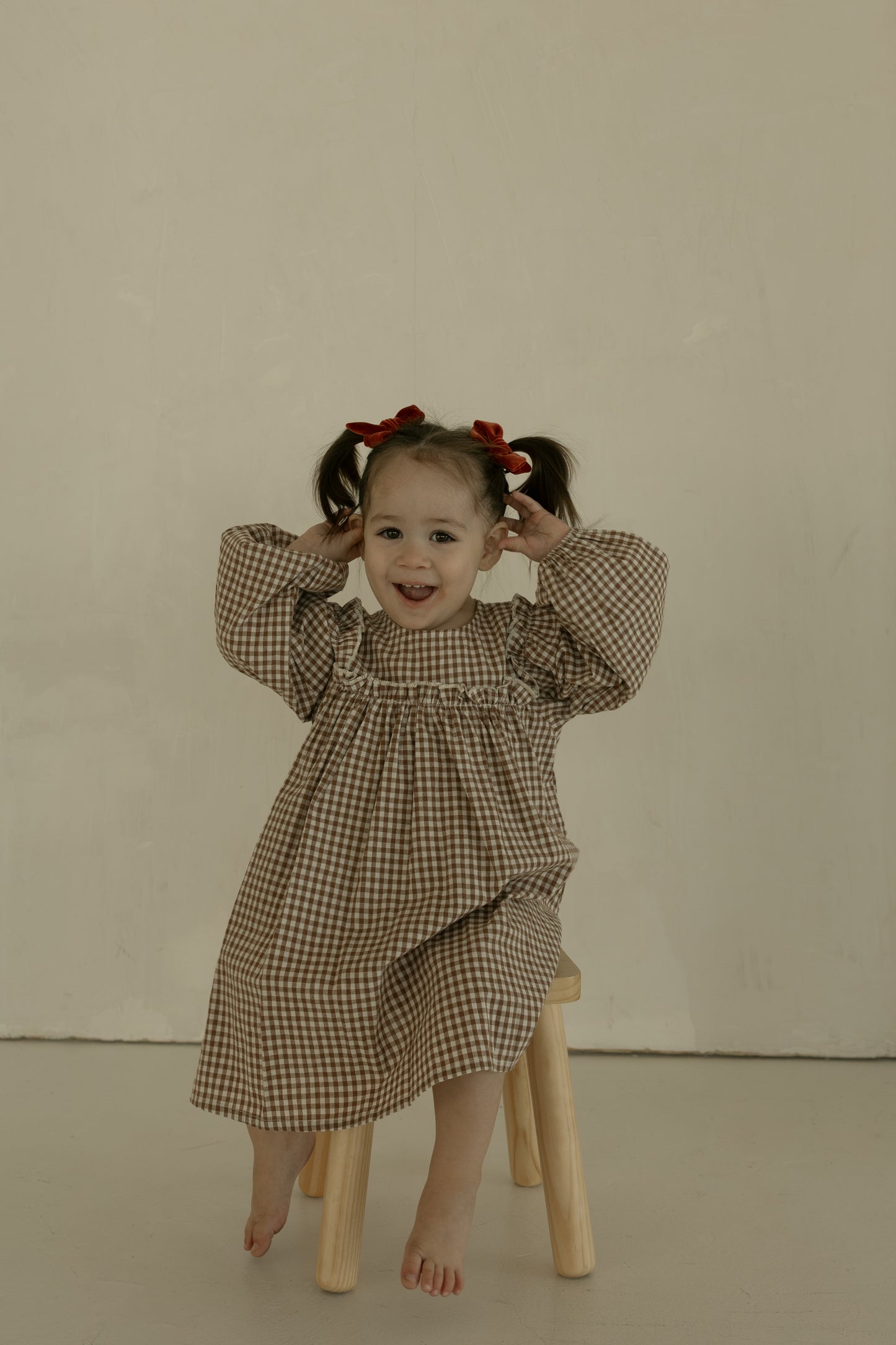 A young child with pigtails tied with red bows sits barefoot on a small wooden stool, smiling and holding her hands near her head, wearing the forever french baby Child Long Sleeve Dress | Honey Gingham against a plain background.