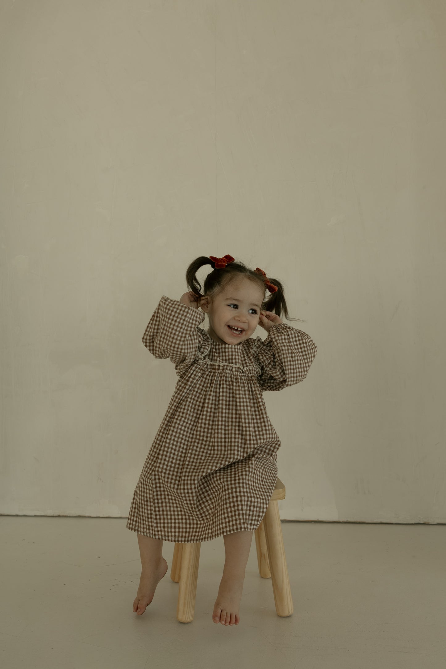 A young girl with pigtails and red bows sits barefoot on a stool, smiling and posing in the forever french baby Child Long Sleeve Dress | Honey Gingham against a plain, light background.