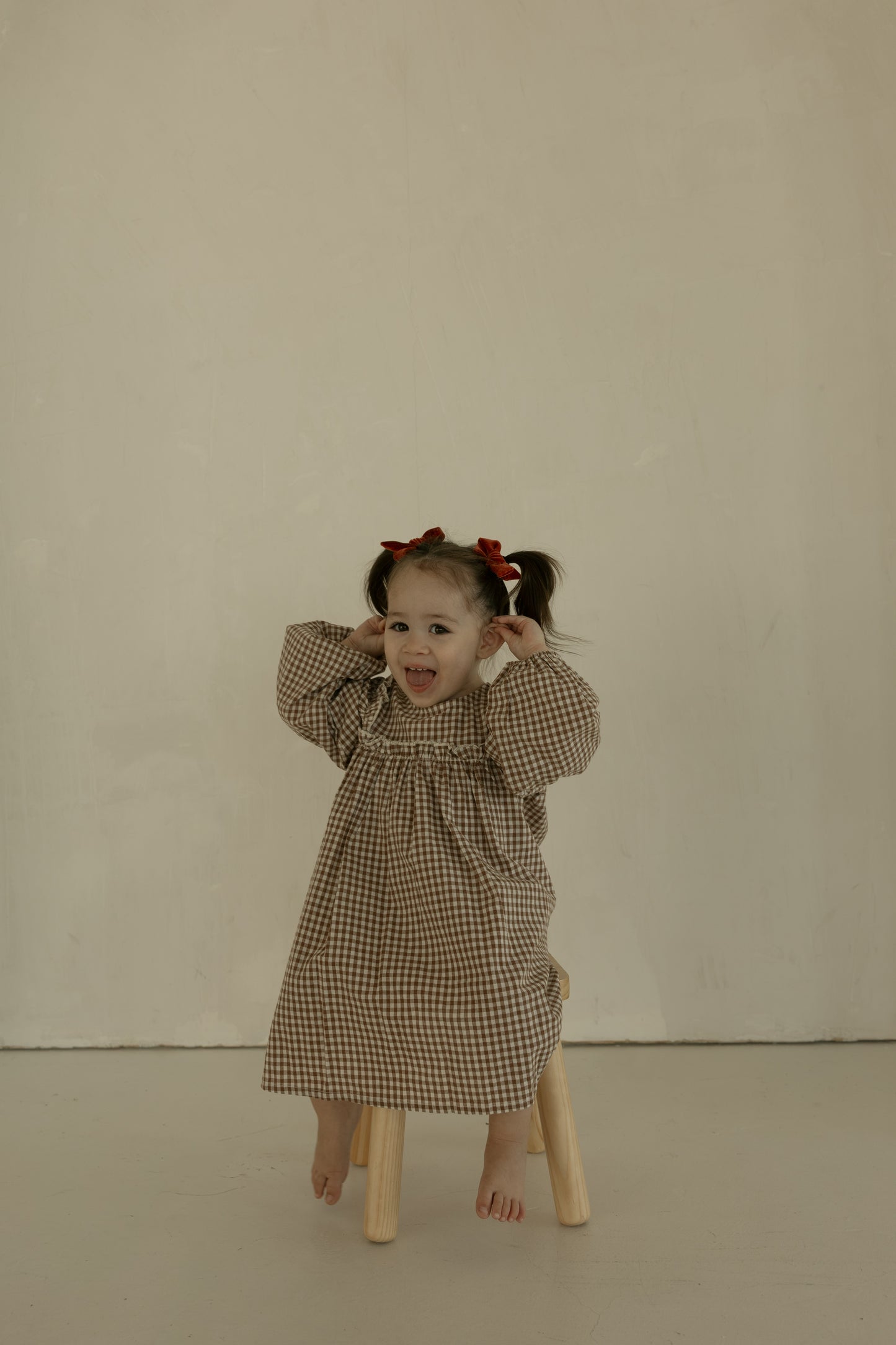 A young girl with pigtails and red bows sits barefoot on a stool, smiling playfully in the forever french baby Child Long Sleeve Dress | Honey Gingham against a plain beige background.