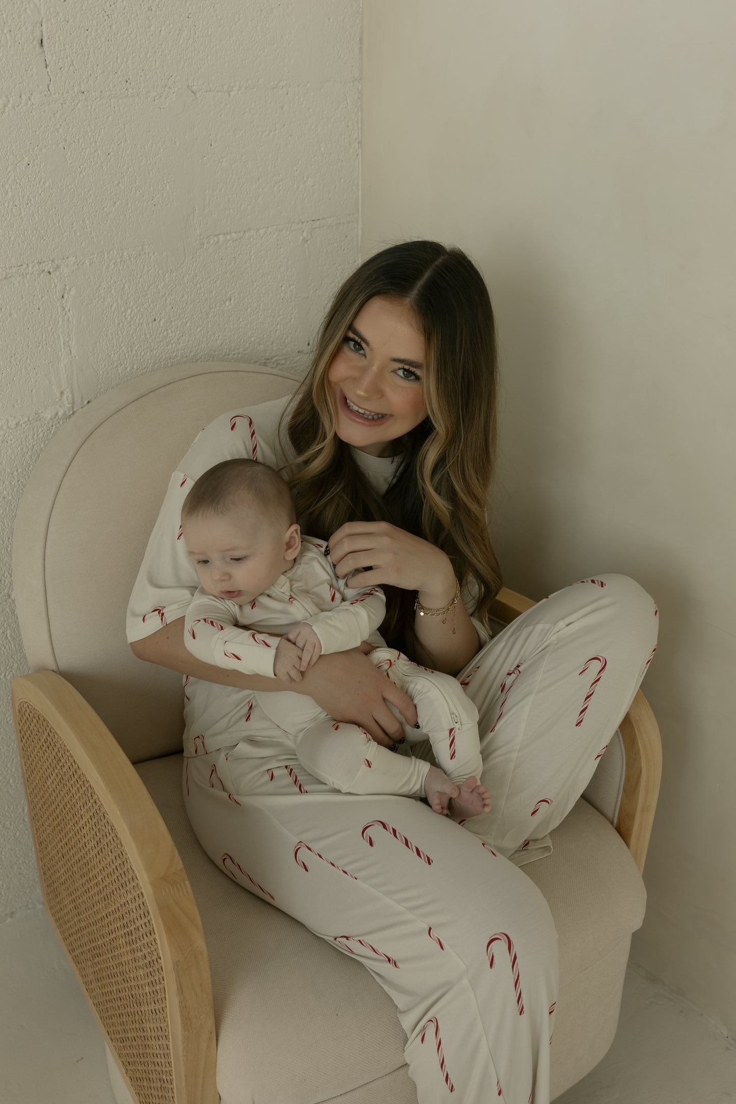 A woman wearing forever french baby Bamboo Zip Pajamas | Candy Cane sits on a beige chair, holding a smiling baby in matching hypo-allergenic pajamas. Both are happy together in a softly lit, minimal room.