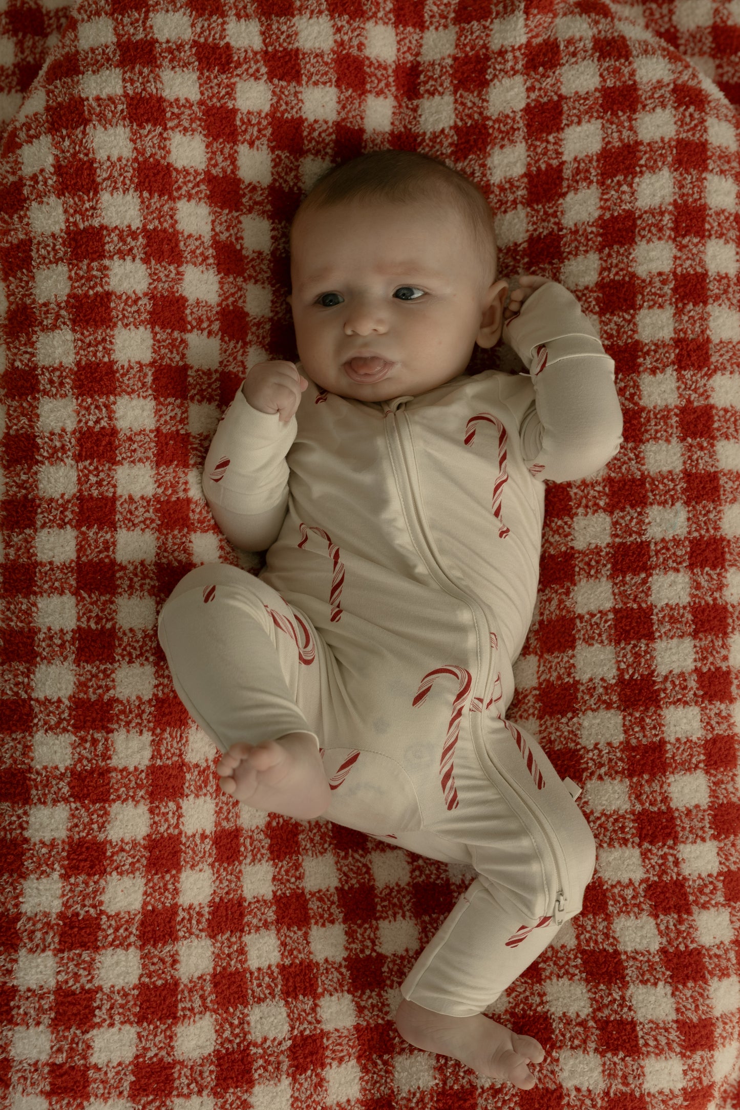 A baby in forever french baby's Bamboo Zip Pajamas | Candy Cane—a cream onesie with red candy canes—lies on a red and white checkered blanket, looking to the side with one arm raised and legs bent.