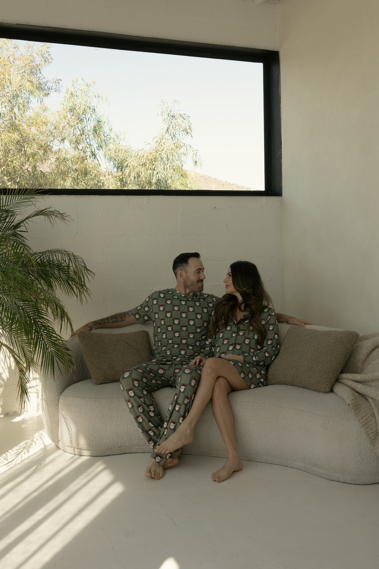A man and woman sit closely on a light sofa in a bright room with a large window, both wearing forever french baby hypoallergenic bamboo pajamas—the man in the Men's Short Sleeve Bamboo Pajamas | Santa Checkerboard.